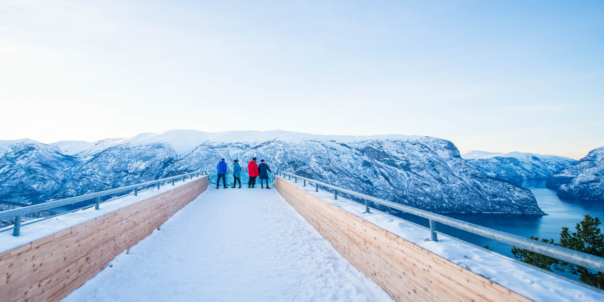 Fire personer på Stegastein utsiktpunkt. Utsikt til Aurlandsfjorden og snøkledde fjell. 