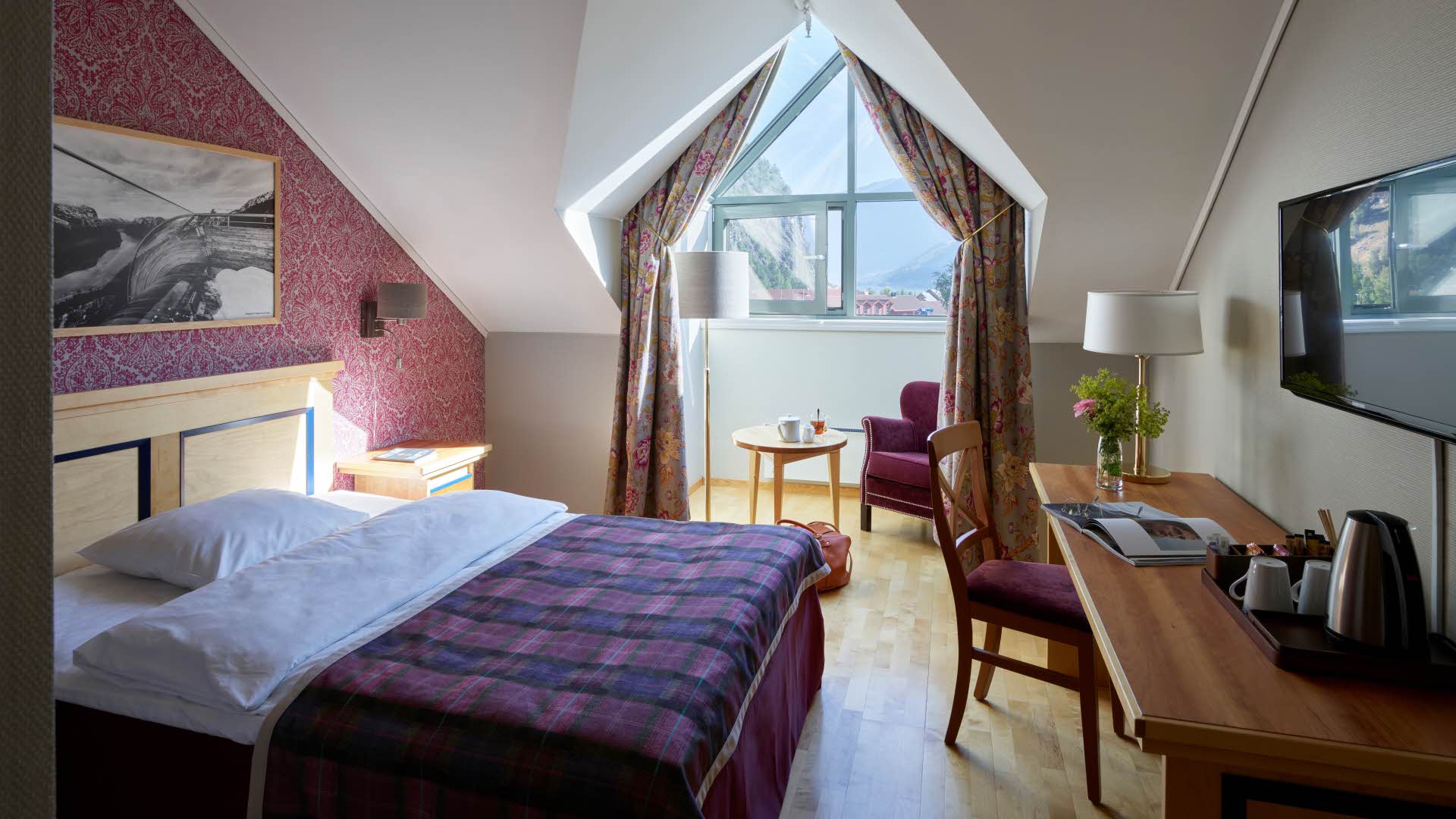 Single room at Fretheim with a queen size bed and desk in front and a chair and table in a bay window in the back. 