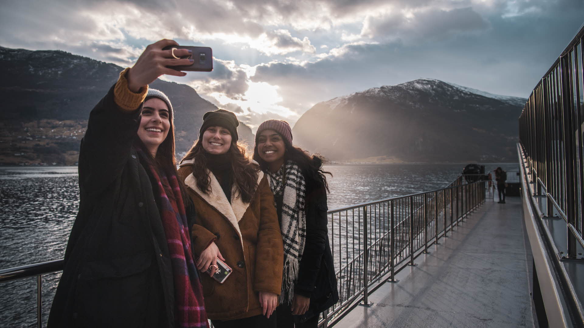 Drei Freundinnen machen im Winter an Bord der „Future of The Fjords“ auf der Fahrt durch den Aurland-Fjord ein fröhliches Selfie