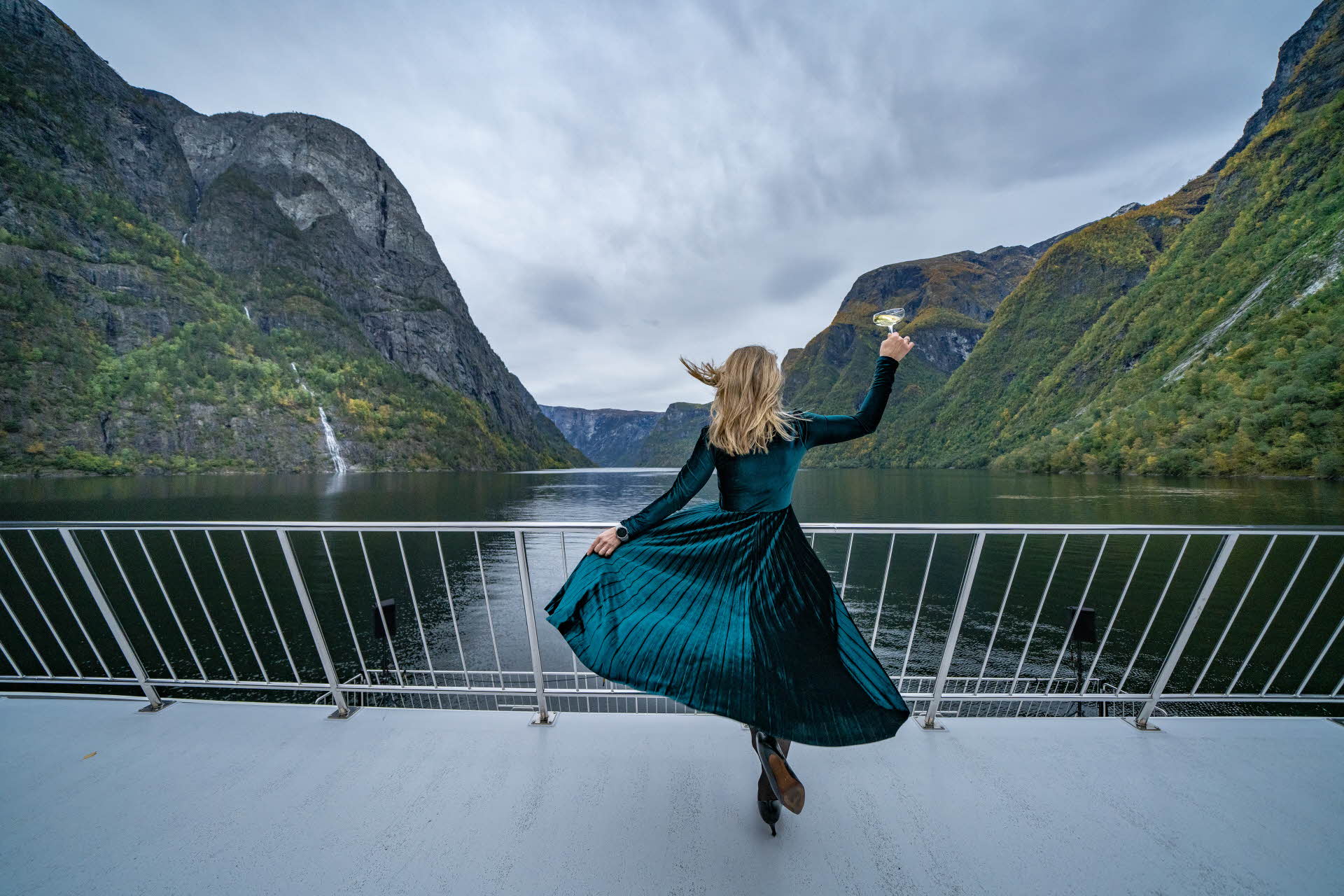 Une femme en robe de velours vert et en talons hauts à l’avant d’un bateau, levant son verre vers le Nærøyfjord.