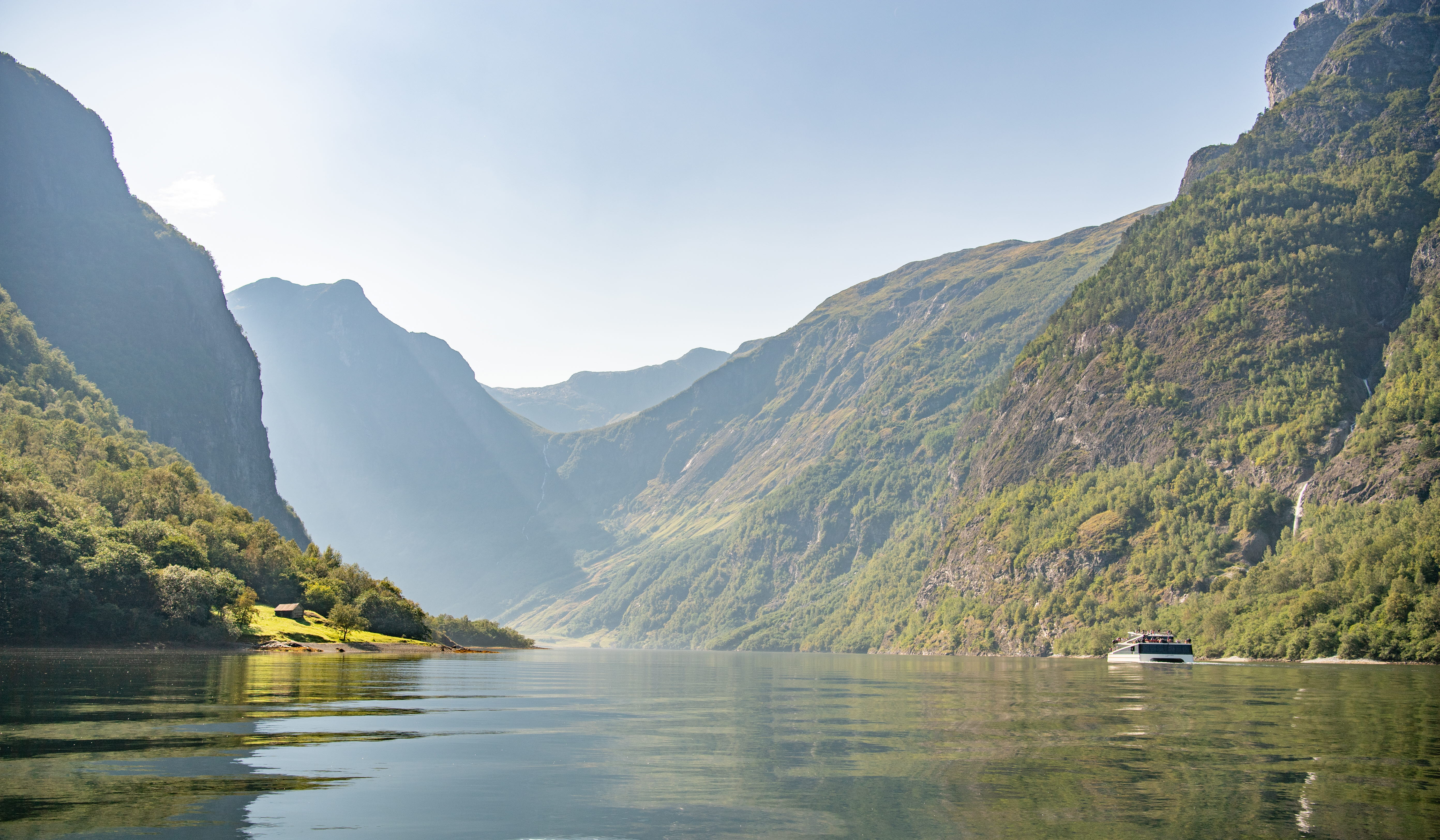 Un bateau électrique à passagers naviguant dans le Nærøyfjord, devant une plaine verdoyante et entre des montagnes escarpées et tout aussi verdoyantes
