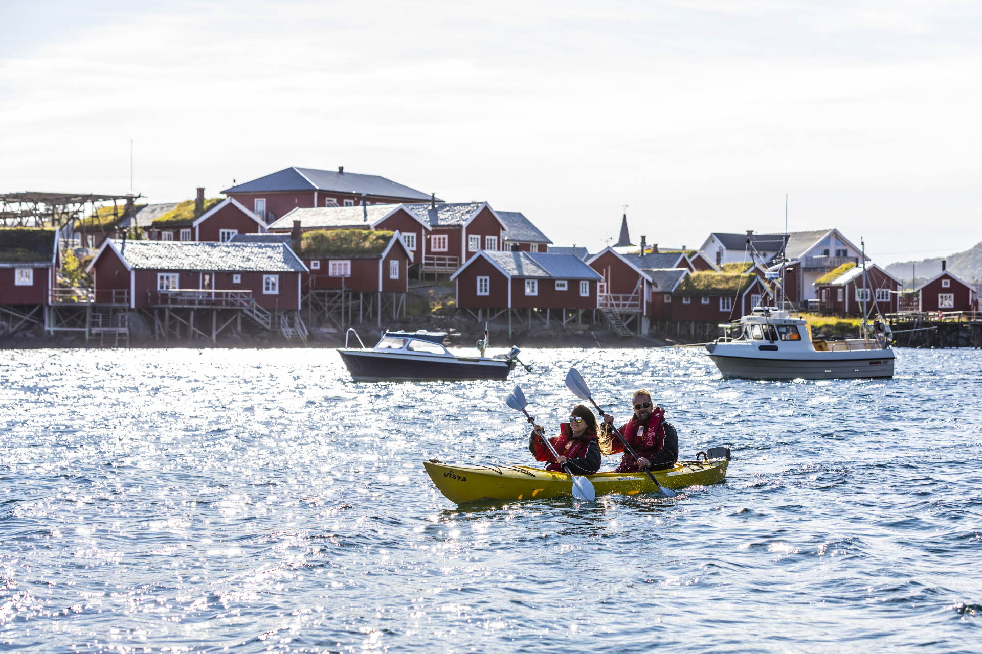 To personer i tandemkajakk på sjøen foran to små båter og Reine Rorbuanlegg.