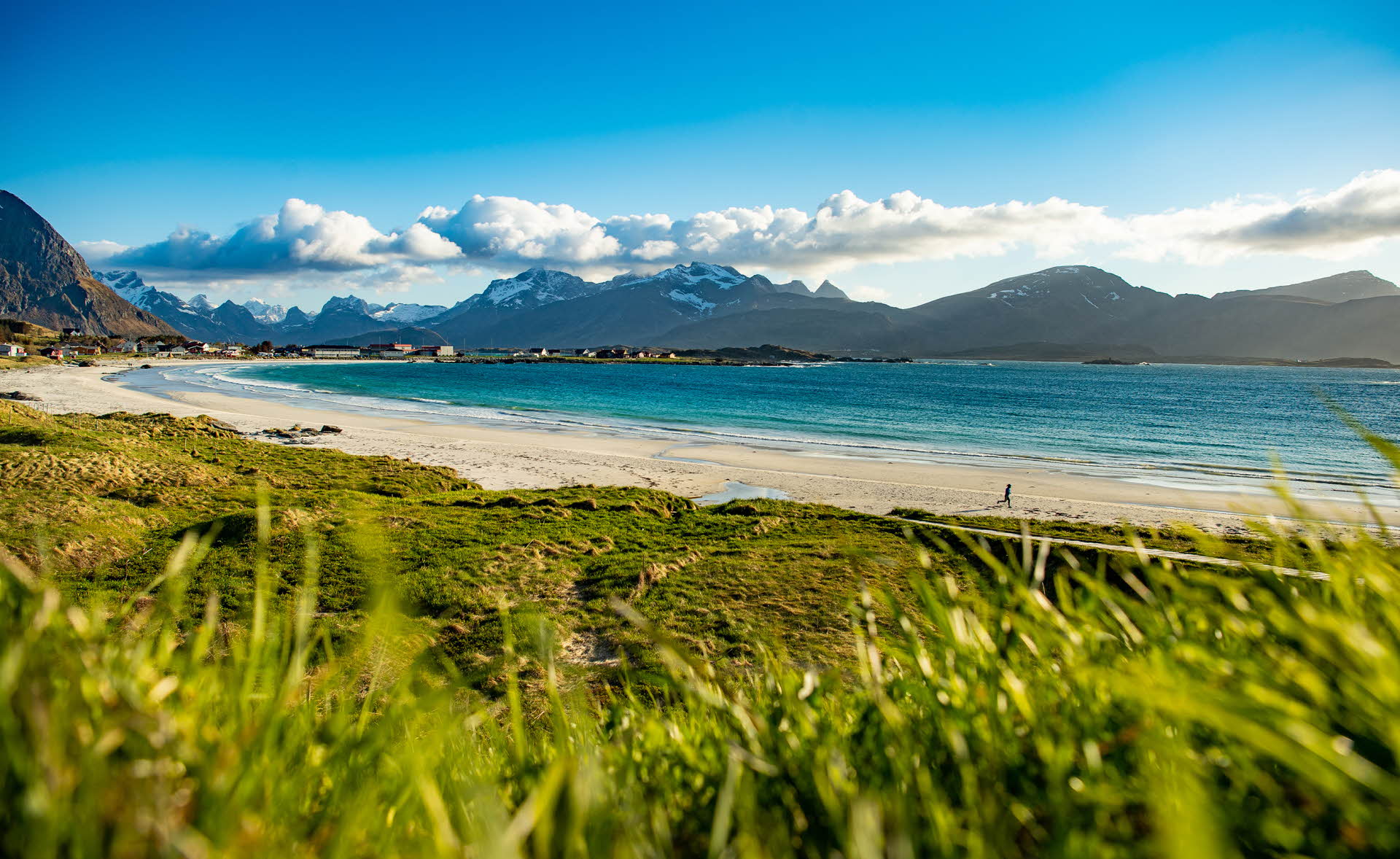 Green grass in front of a white sandy beach, blue sea and dark mountains in the background.