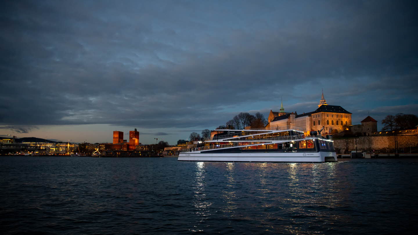 Legacy of The Fjords on Oslofjord, passing Akershus Fortress late at night with lit city line