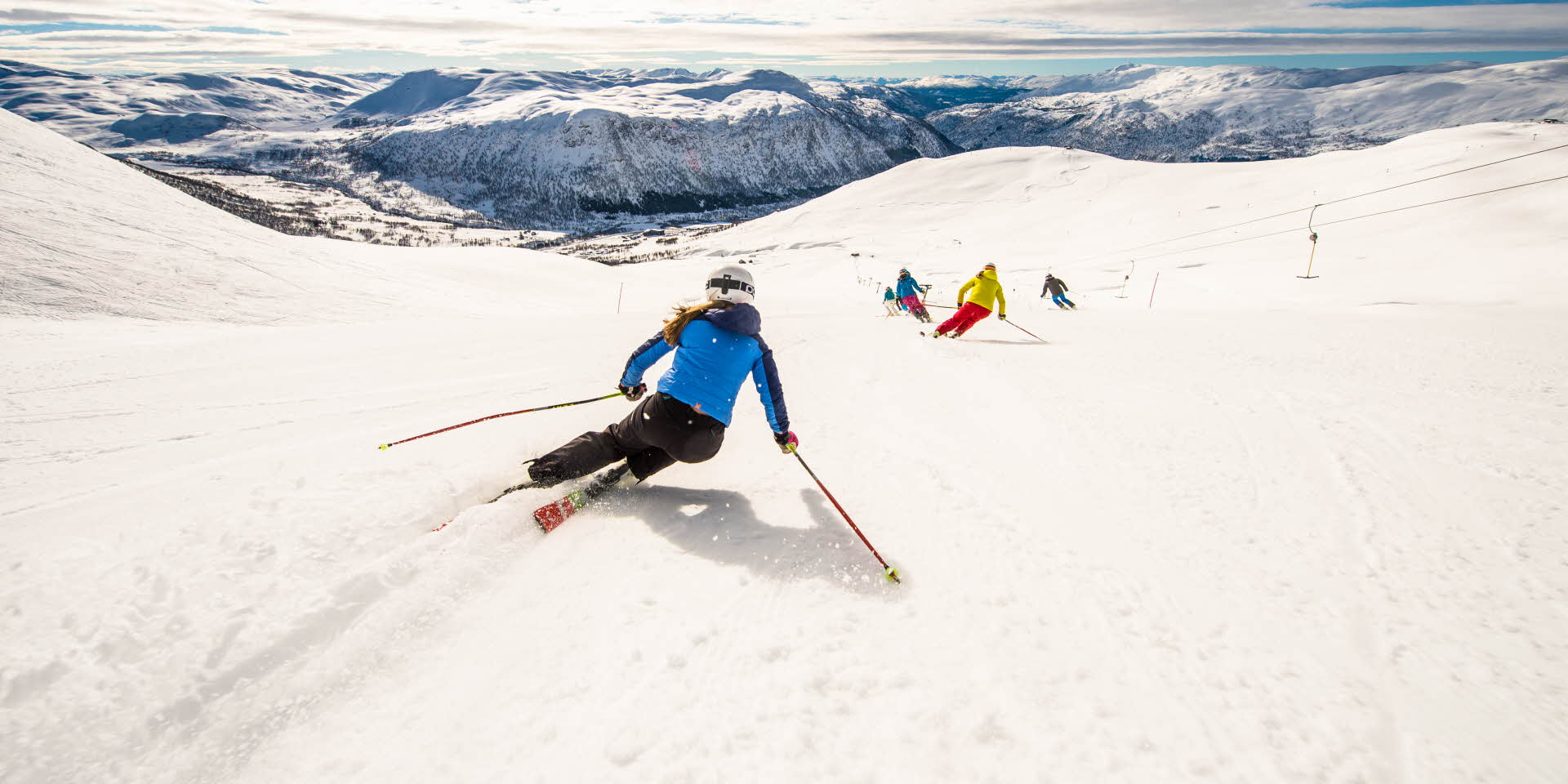 A woman in blue jacket and white helmet skiing behind other people on a slope with views to Myrkdalen. 