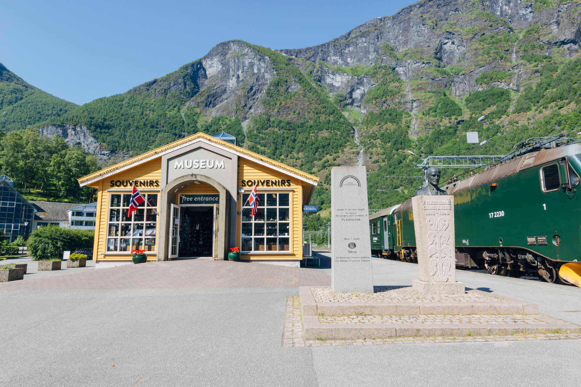 The exterior of Flåm Railway Museum next to a green train. A sculture in front and tall mountains with green forest behind. 