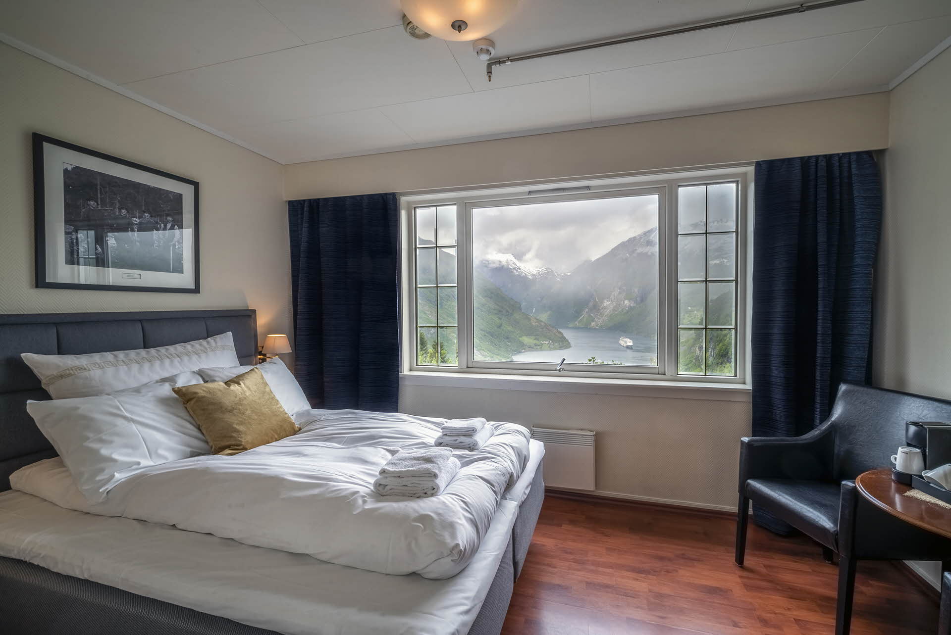 Interior of a double room at Hotel Utsikten. Bed, lounge chair, table and window with fjord view. 