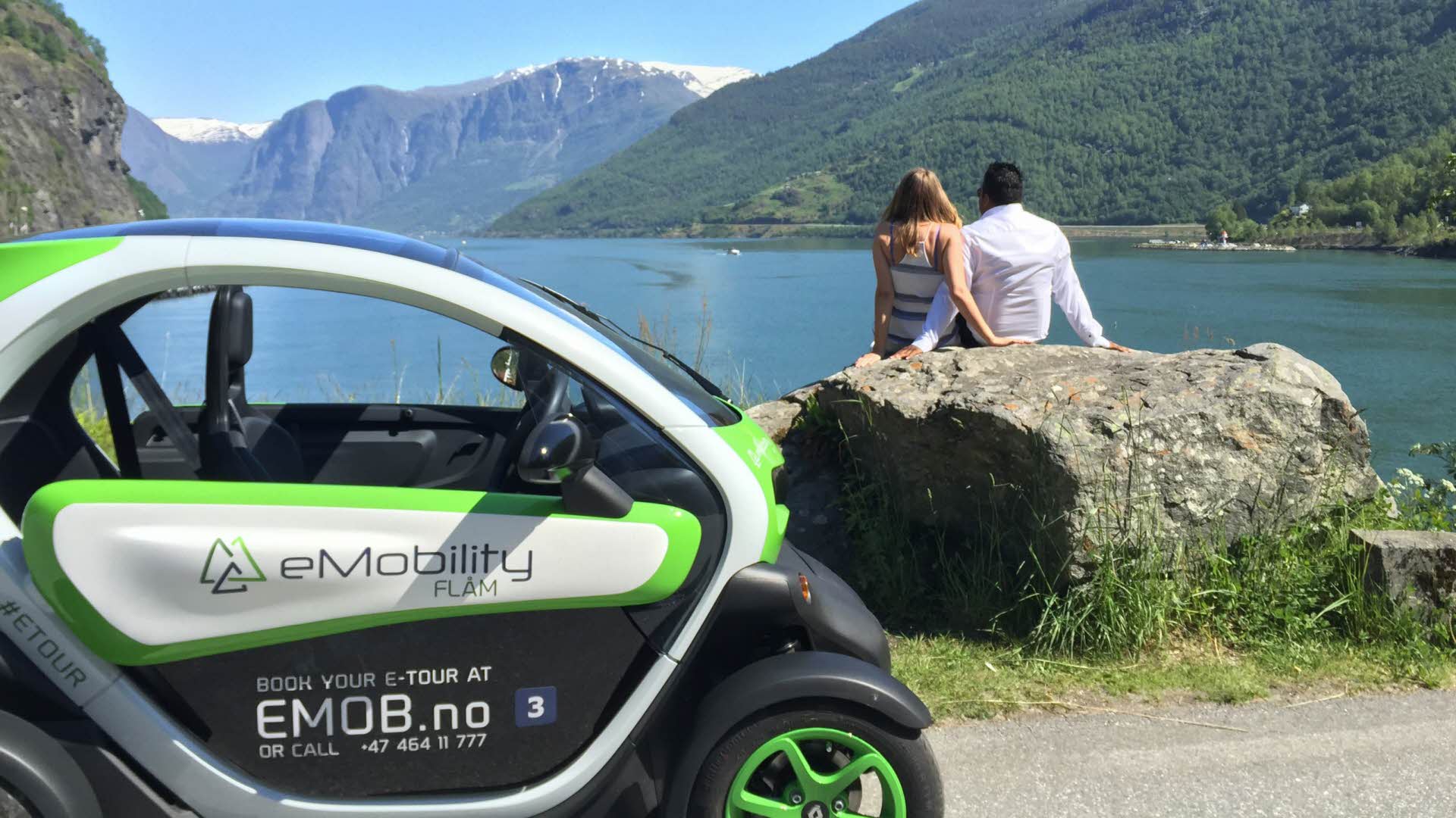 Una pareja sentada en una roca junto al mar mirando al fiordo y a la montaña, con un coche eléctrico al fondo