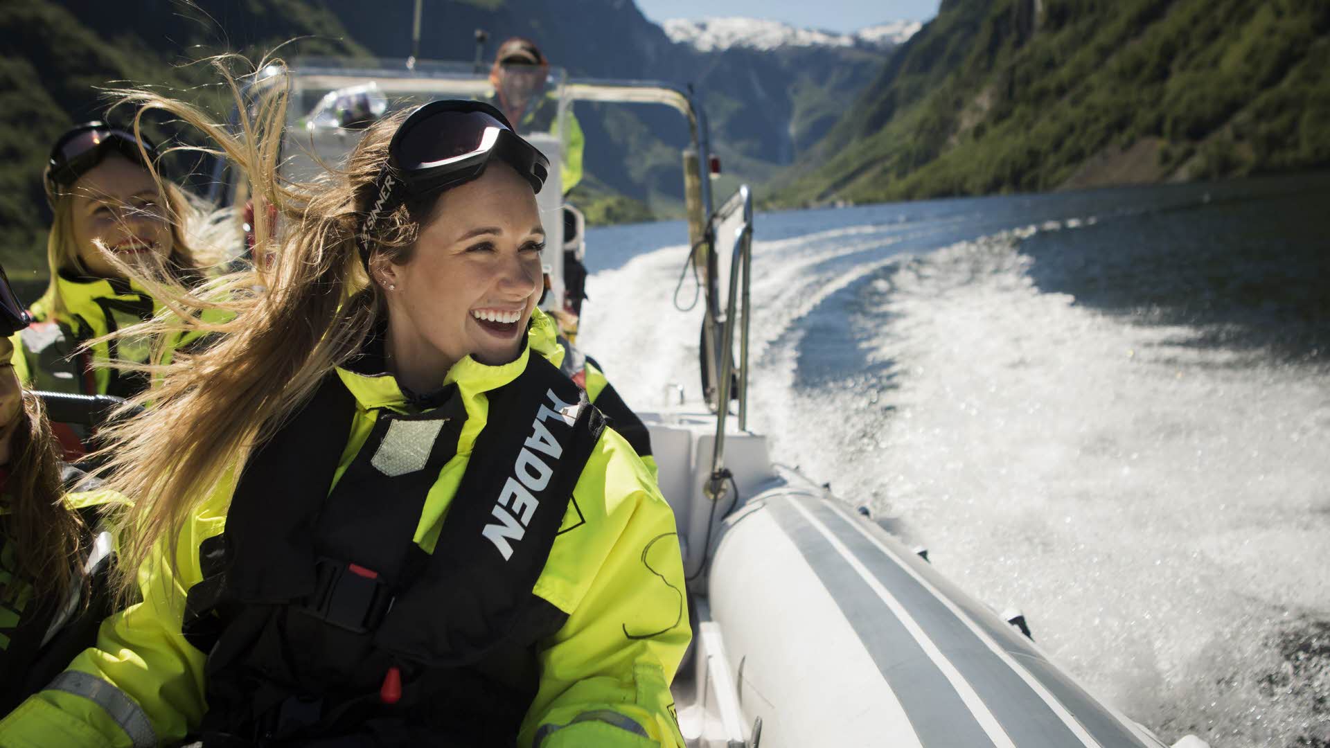 Une femme souriante avec un gilet de sauvetage et des lunettes sur la tête est assise dans un bateau semi-rigide qui fait des vagues dans le fjord. 