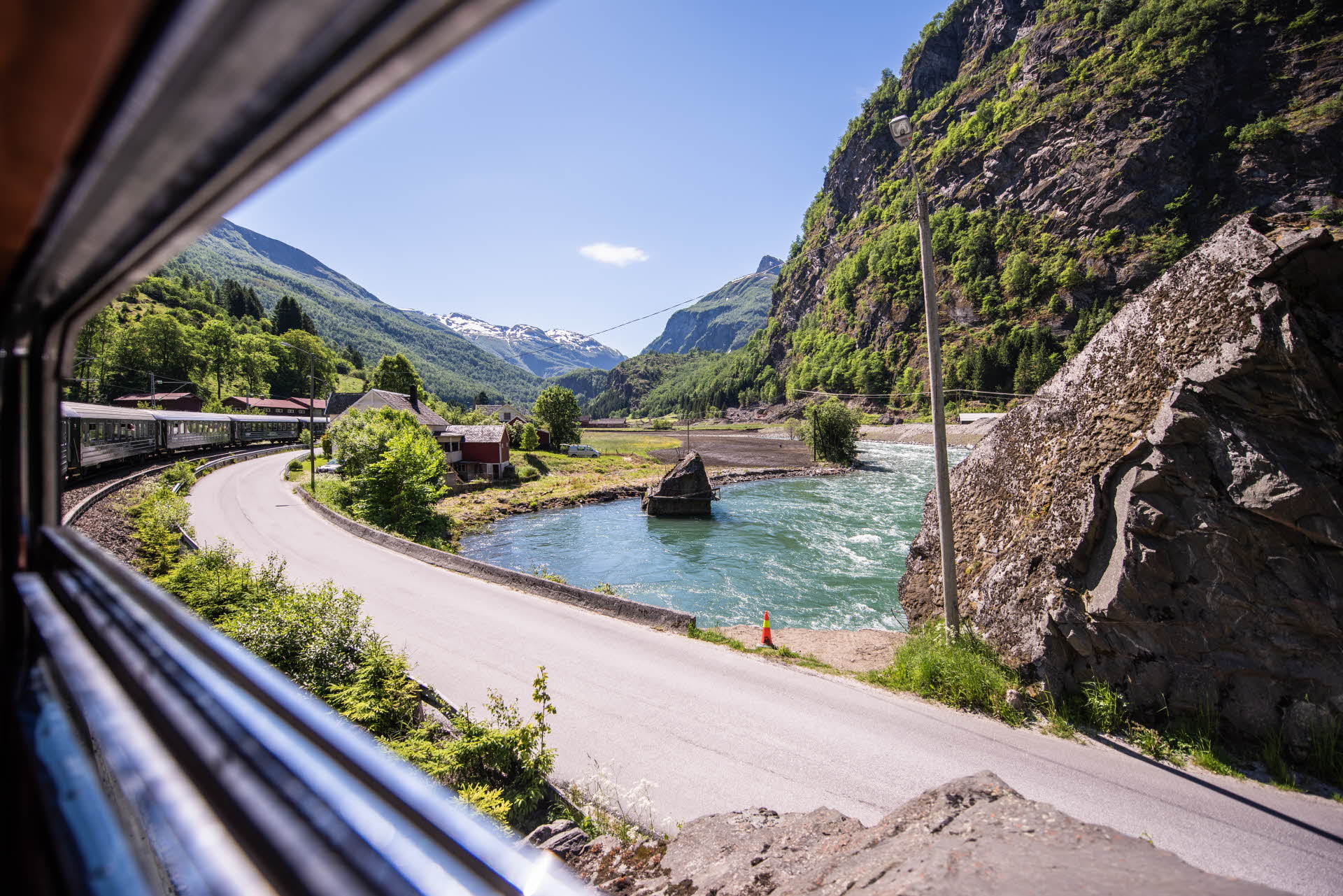 Vue depuis l’intérieur de la Flåmsbana alors que le train traverse la rivière dans un virage parallèle à la route, sur fond de montagnes luxuriantes