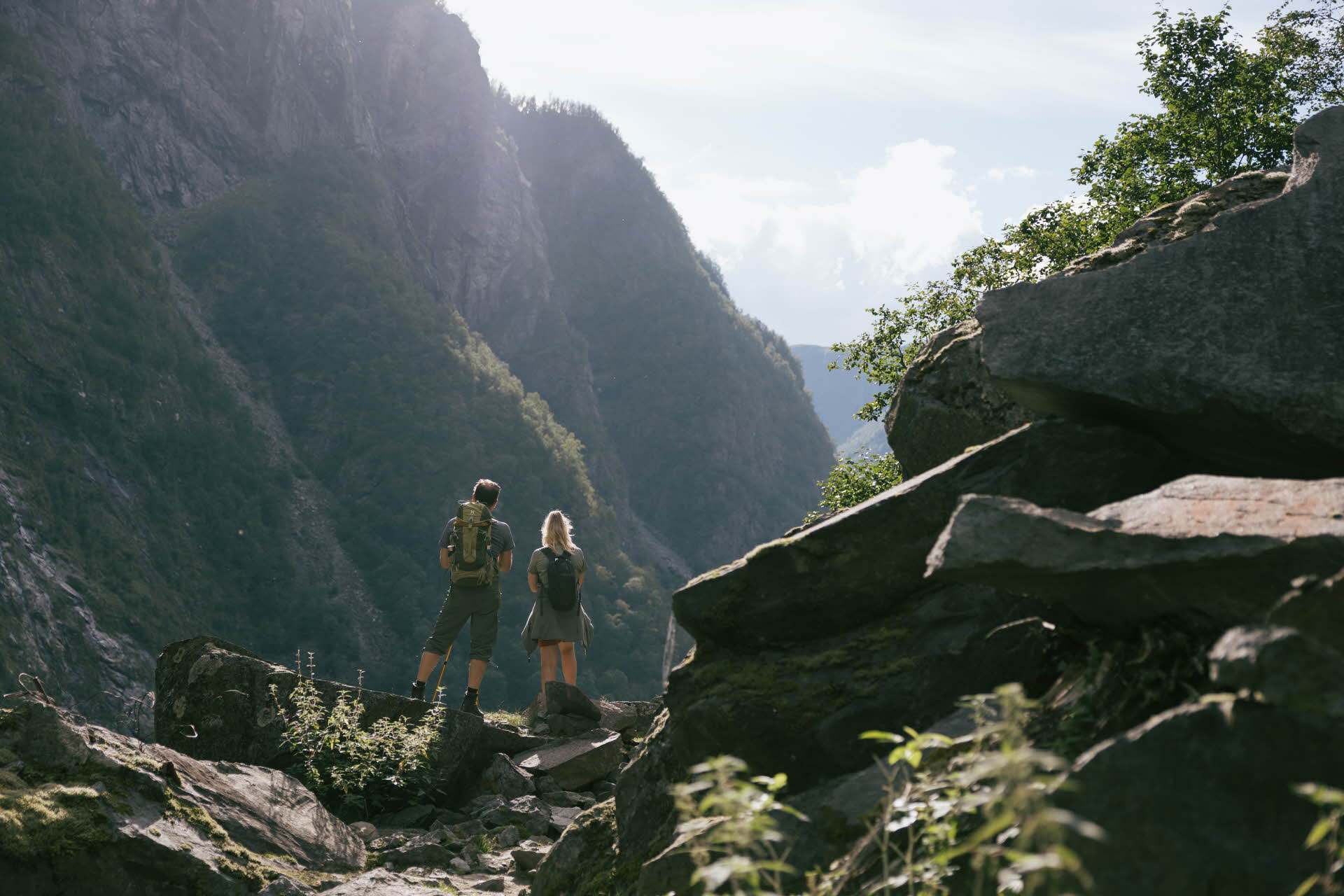 To personer står med ryggen til mellom store kampesteiner og ser mot de bratte fjellsidene i Aurlandsdalen