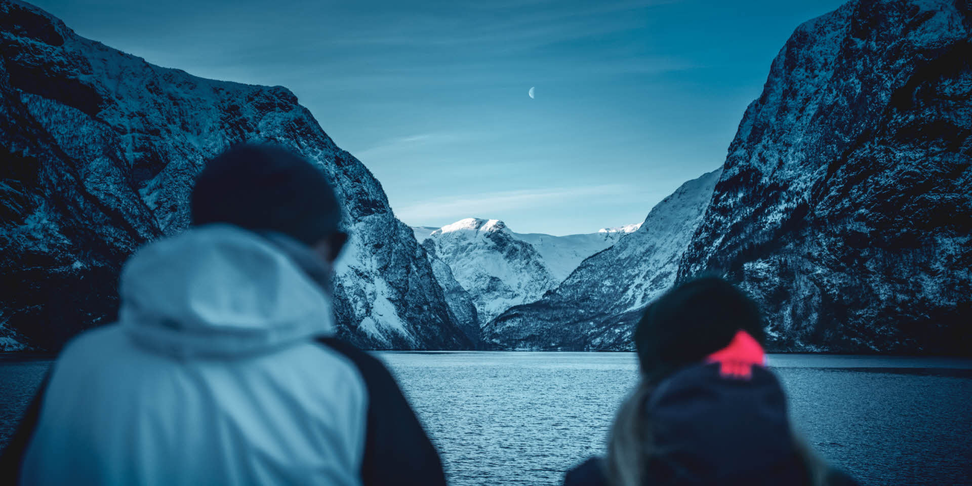 To personer står på dekk på Future of The Fjords og ser ut over Nærøyfjorden og snødekte fjell.