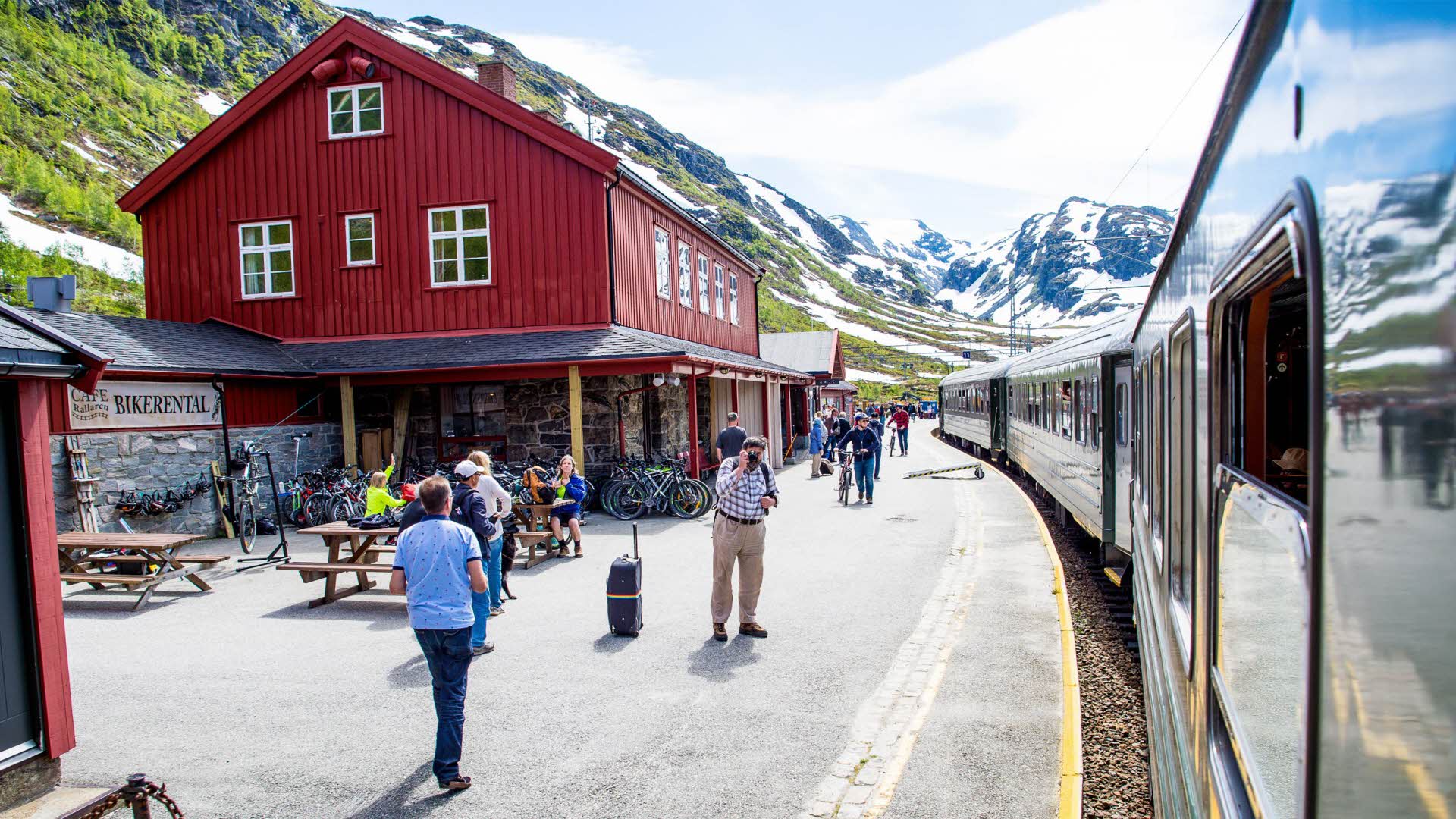 Tourists outside the Flåm railway at Myrdal station. Red station building and snow-patched mountains. 