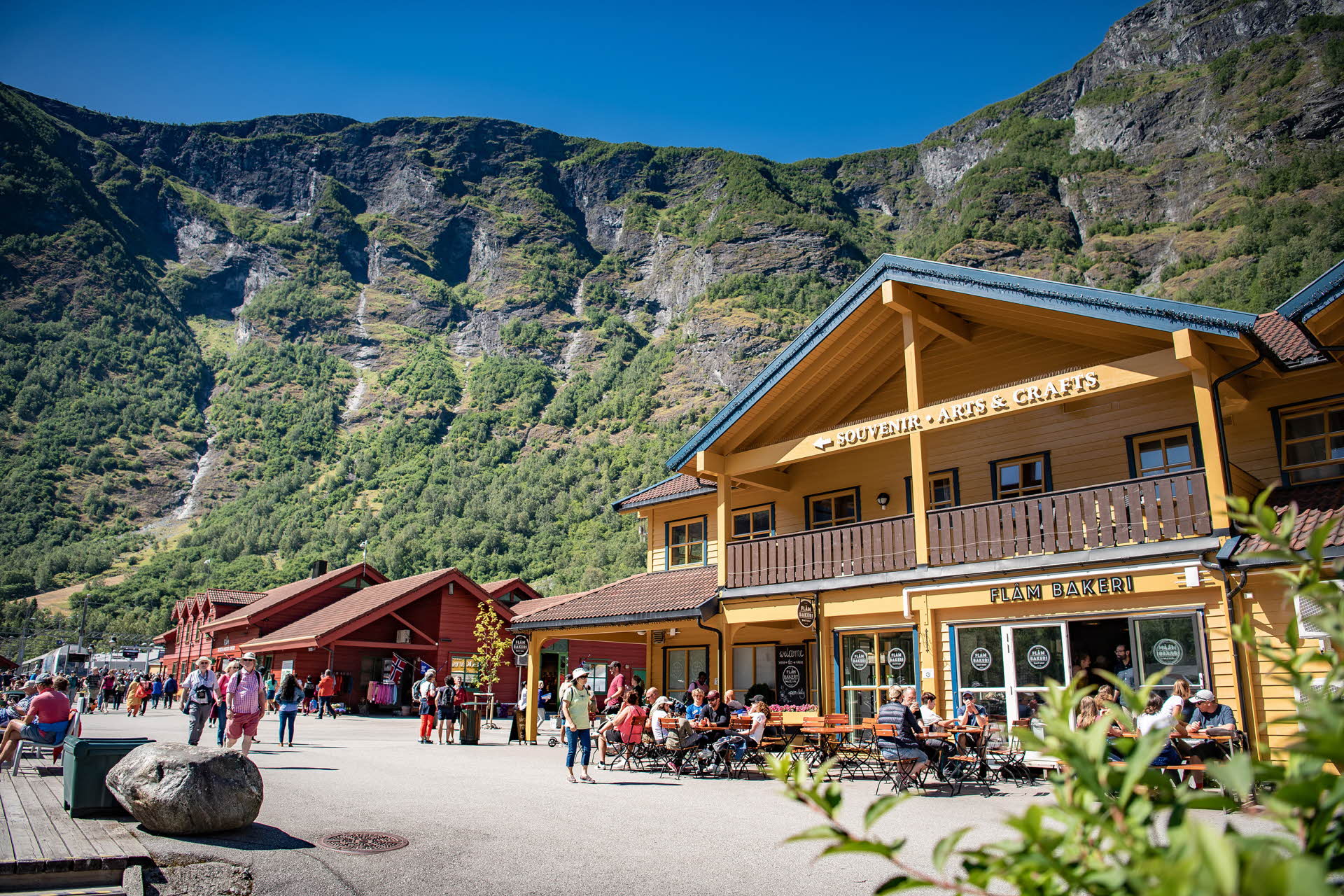 Un bâtiment jaune avec des tables disposées à l’extérieur de la Flåm Bakeri. Des touristes dans la rue, une maison rouge et de hautes montagnes. 