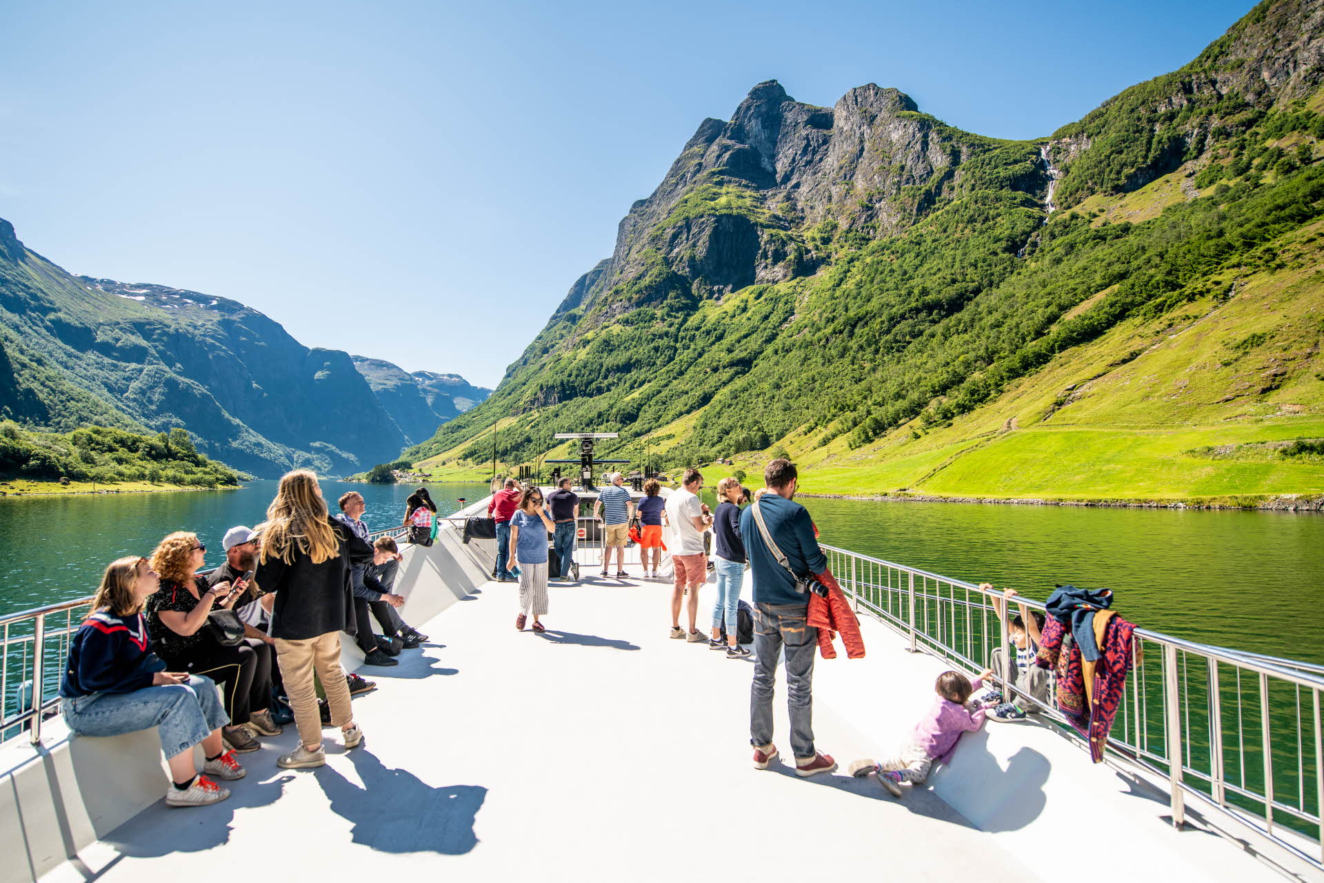 People sitting and standing on the roof of a boat in the Nærøyfjord, with views towards green mountainsides