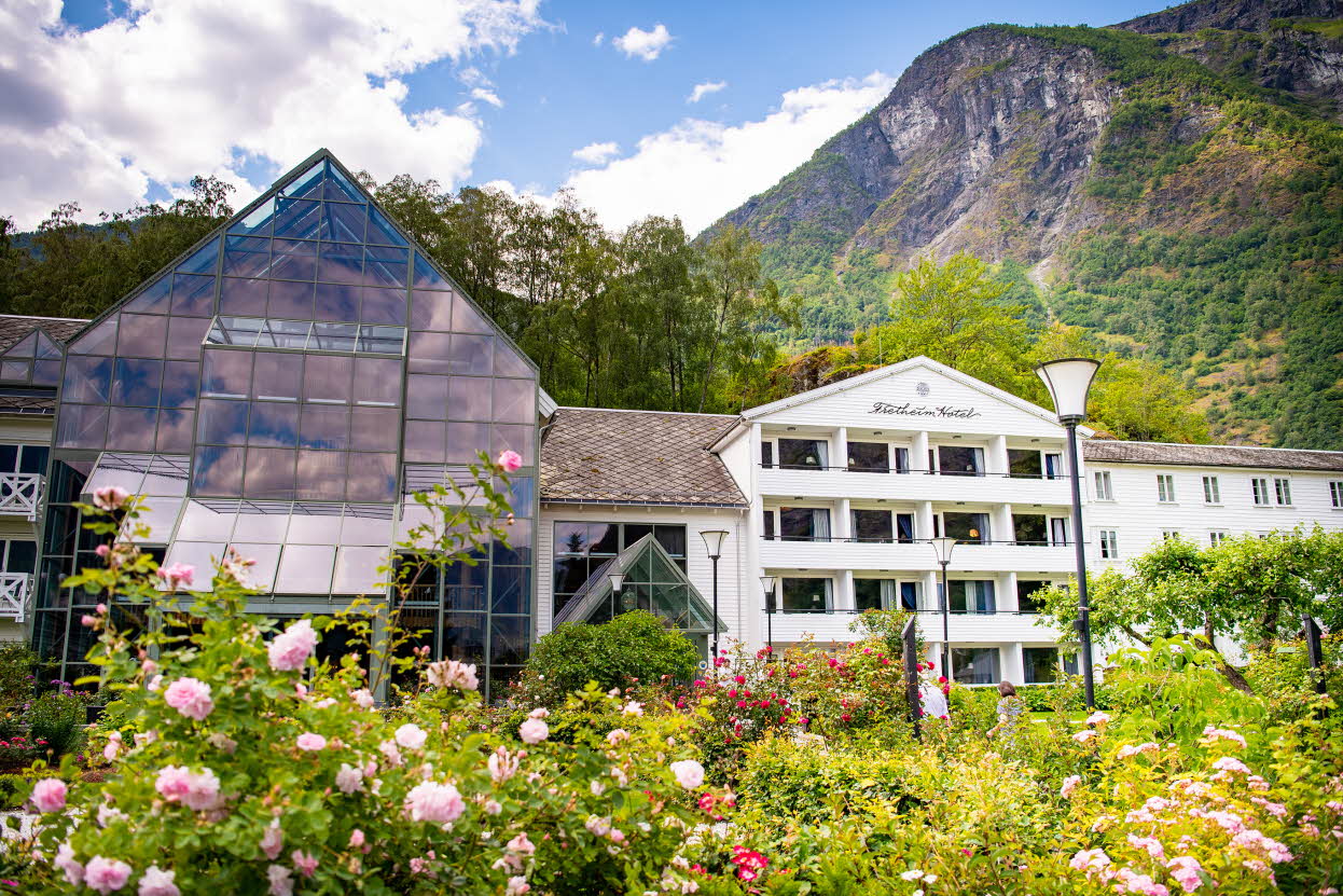Fretheim Hotel sett bak frodige rosebusker. Skog, fjell og blå himmel bak.
