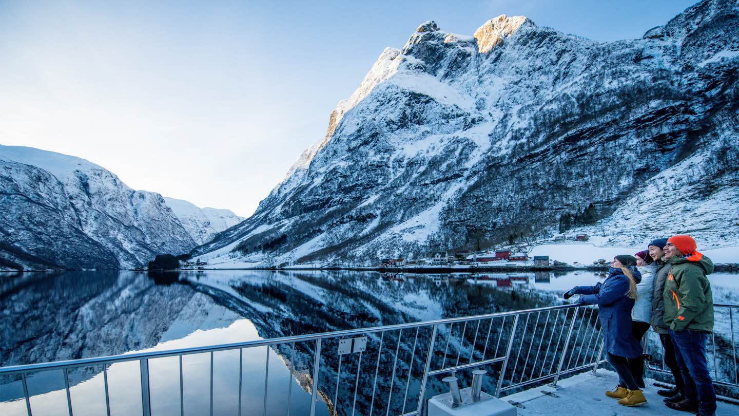 2 young couples front deck of Vision of The Fjords sailing through Naeroyfjord snowy mountains is mirrored in sea