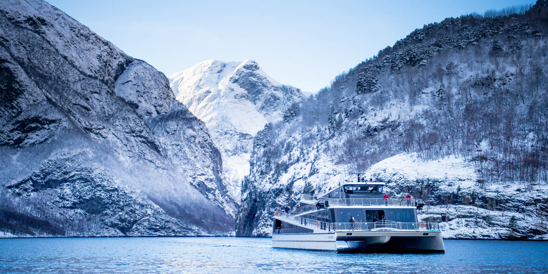 Moderne båt seiler på Nærøyfjorden på vinteren omringet av snøkledde fjell.
