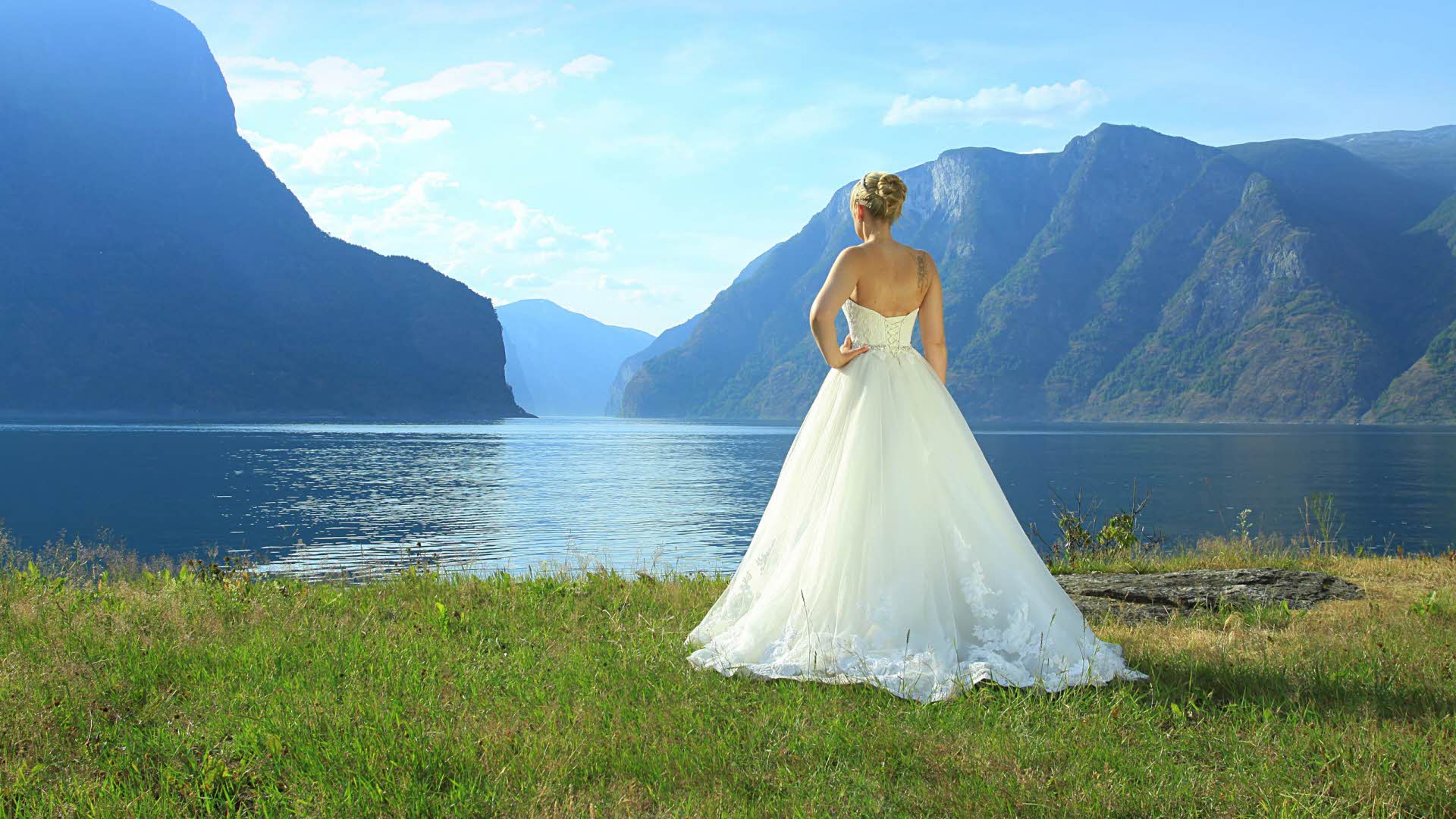 A woman in a long, white wedding dress seen from behind looking out on the Aurlandsfjord.