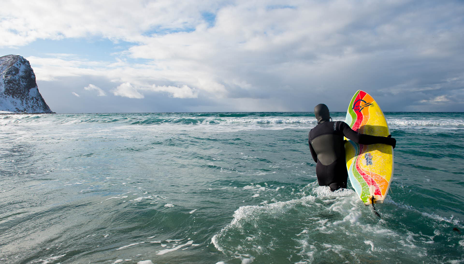 A surfer in a dry suit entering the sea. Mountain to the left.