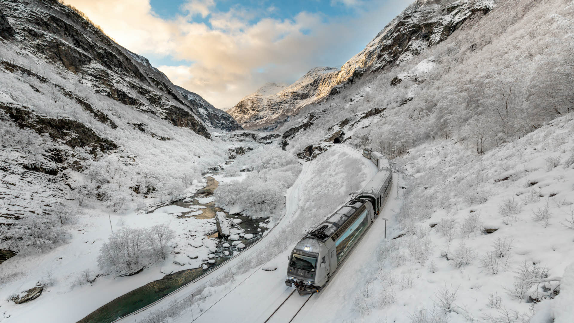 Ein Zug fährt im schneebedeckten Flåmsdalen am Fluss entlang. 