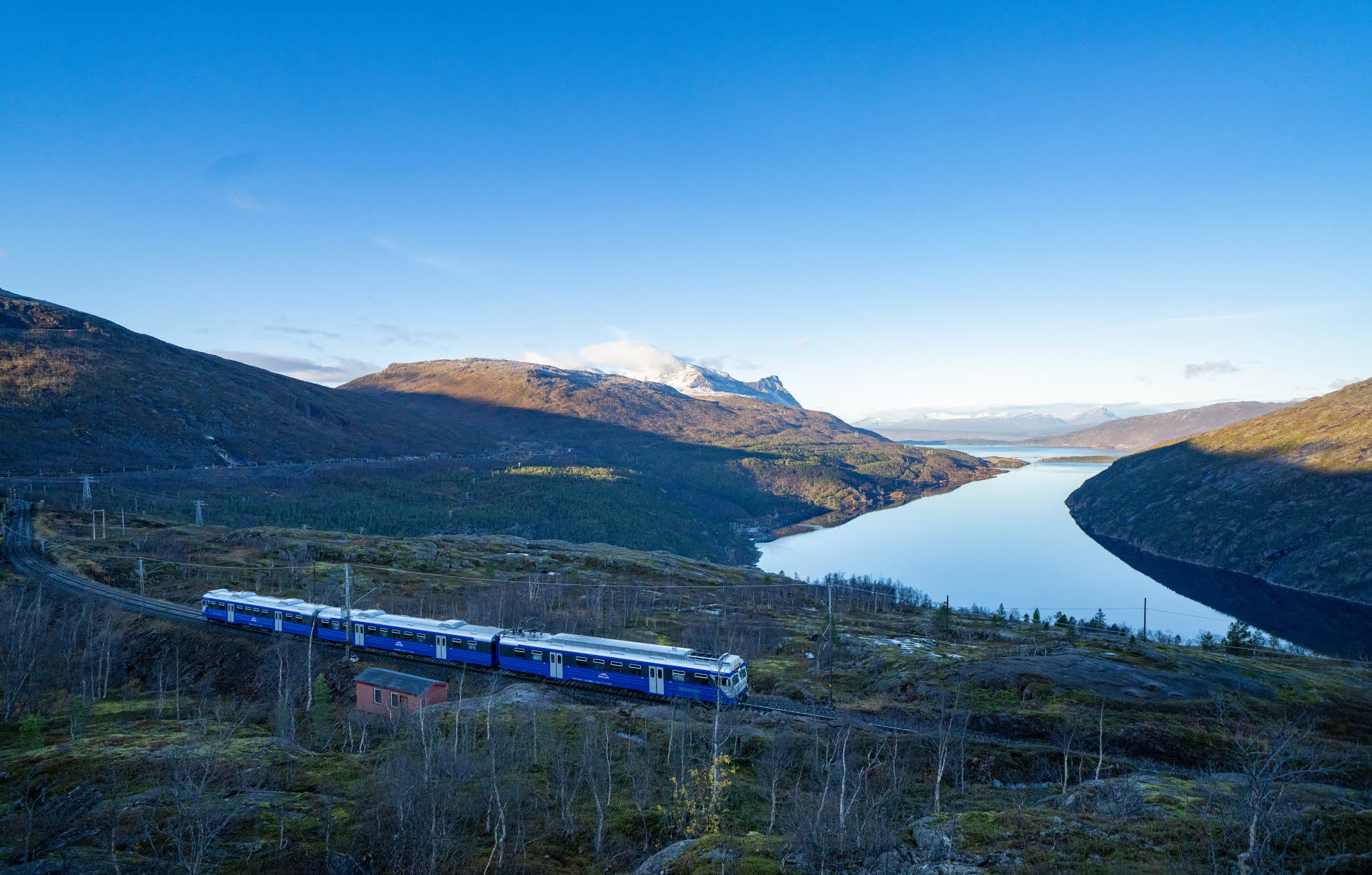El Arctic Train a su paso por una pequeña cabaña roja en el ferrocarril Ofot, en dirección hacia Narvik con el Rombaksfjord y las montañas de fondo