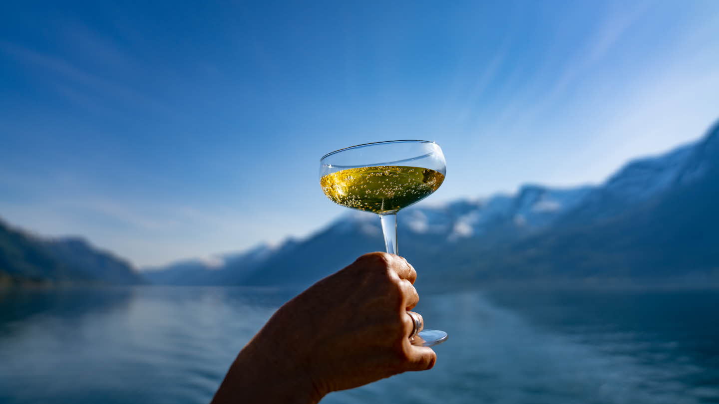 A hand holding a glass with cider. The Hardangerfjord, mountains and blue sky in the background. 