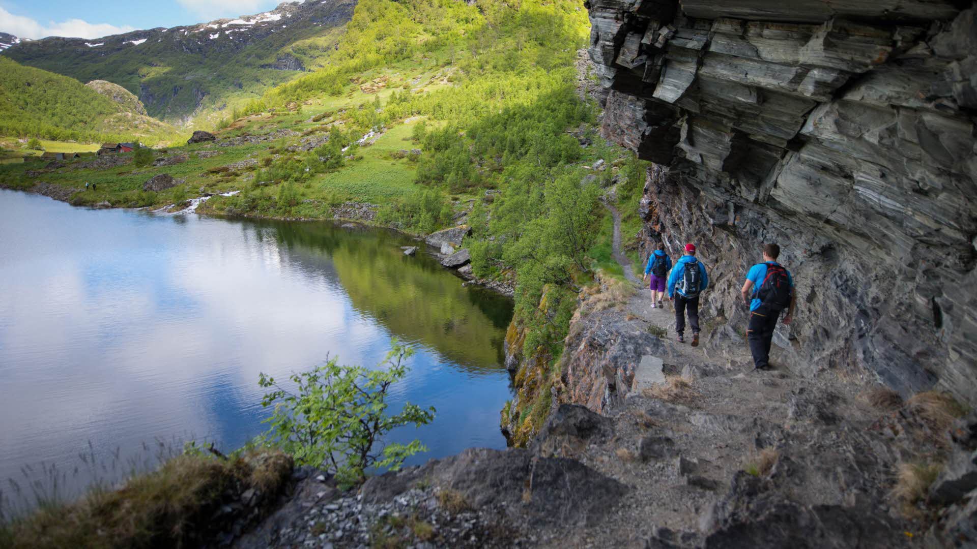 3 turgåere i blå klær går nedover gjennom en bratt fjellhylle ved et stille vann i Aurlandsdalen