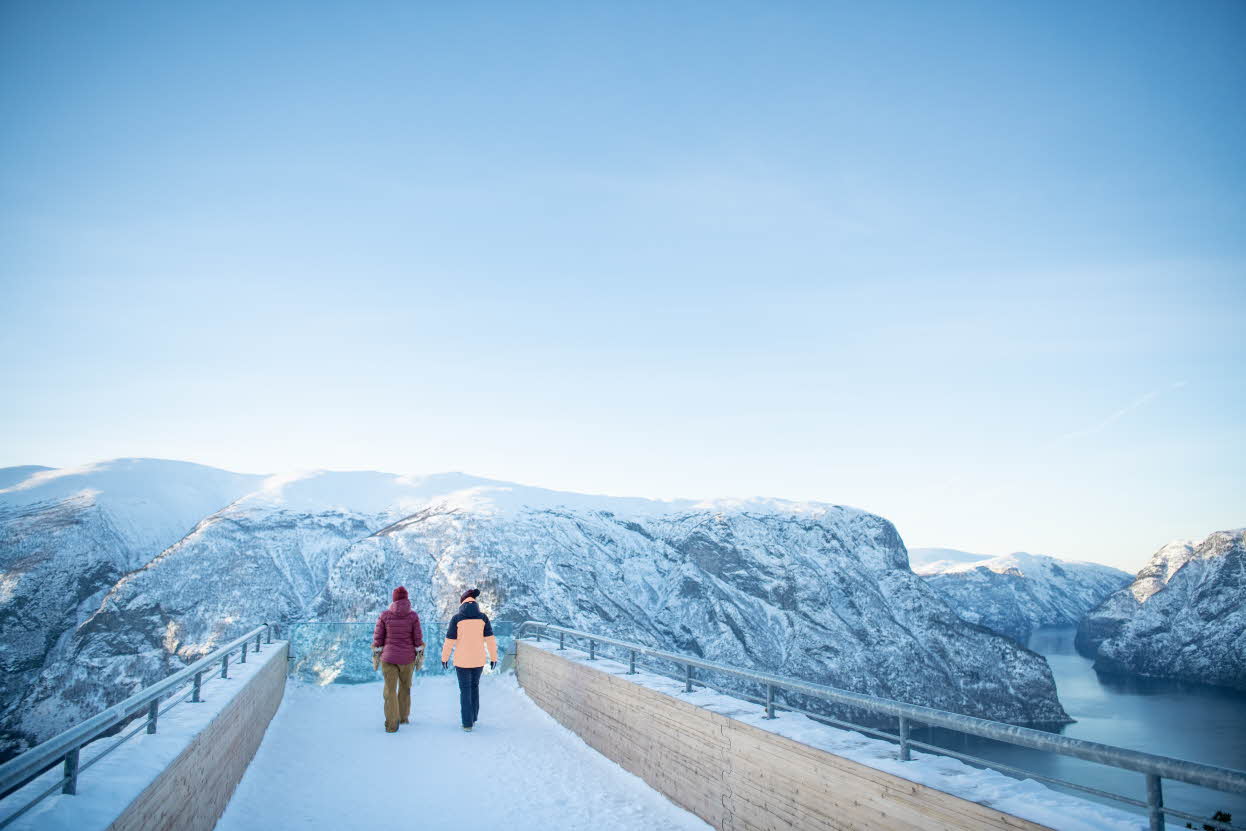 Zwei Menschen laufen zum vorderen Ende der Aussichtsplattform Stegastein. Gedämpftes Licht, verschneite Landschaft und weiter unten der Fjord.