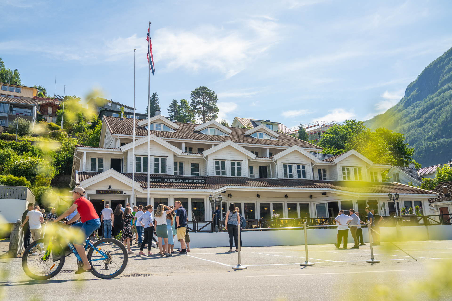 En été, la terrasse extérieure en bois de l’hôtel Aurlandsfjord avec des personnes regroupées devant l’hôtel et un garçon à vélo vêtu d’un T-shirt rouge
