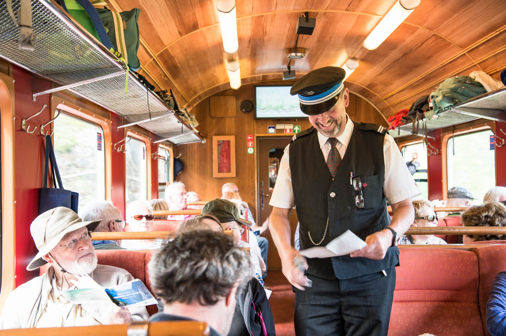 A ticket inspector in uniform and hat smiling while checking a group’s tickets on the Flåmsbana