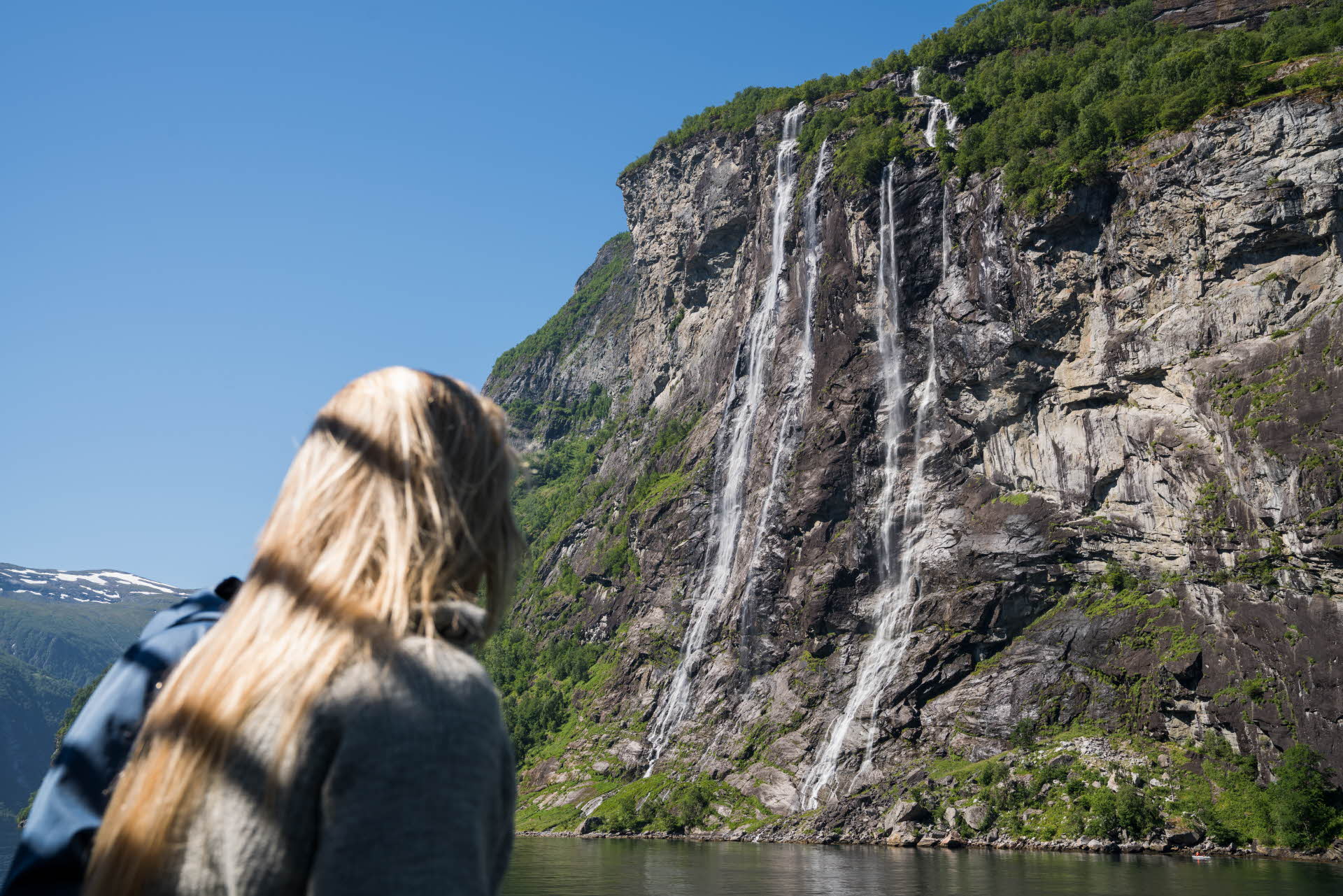 Woman with blond hair onboard Fjord Cruise Geirangerfjord watching huge cascading waterfalls going straight into the fjord