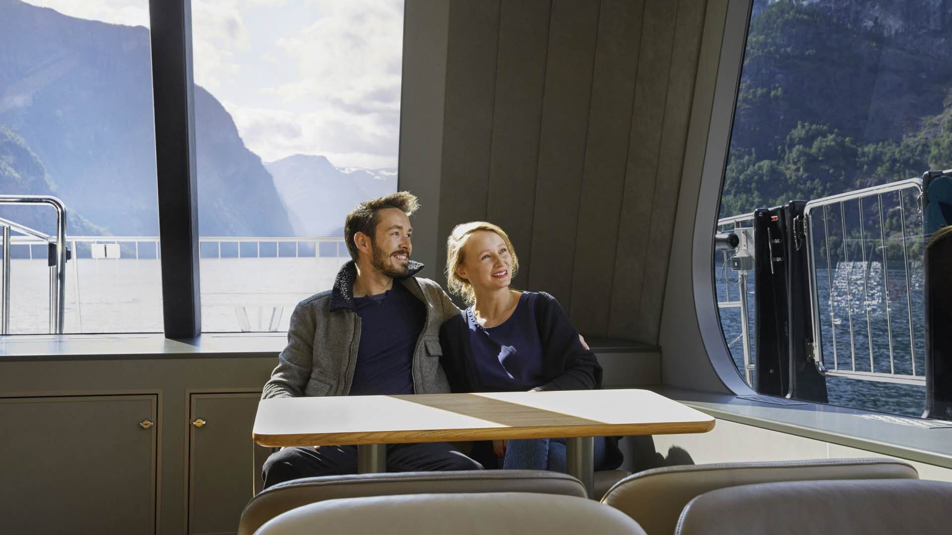 Ein Paar Mitte 20 genießt den Blick auf den Nærøyfjord aus dem Panoramafenster der „Vision of The Fjords“