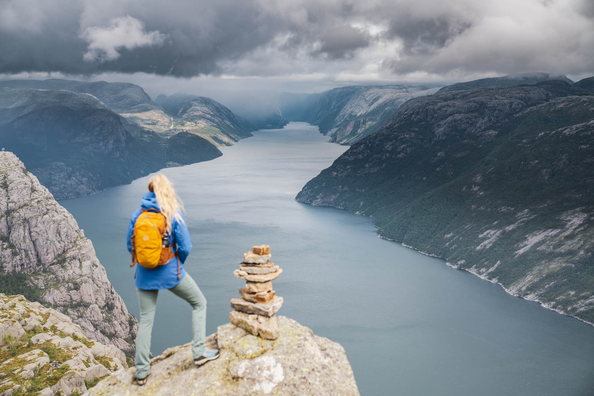 Une femme aux longs cheveux blonds, à la veste bleue et au sac à dos jaune est debout près d’un cairn juste au-dessus du Lysefjord