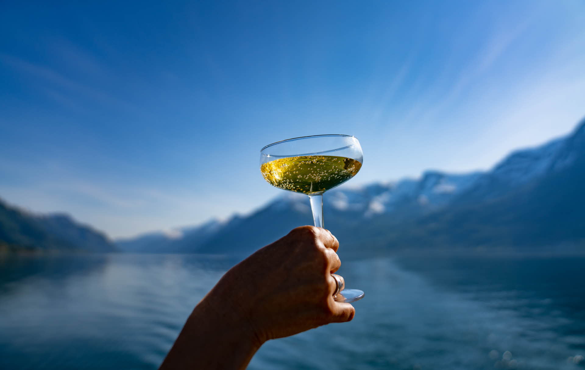 En hånd som holder et champagneglass. Hardangerfjorden og fjellrekker bak. 