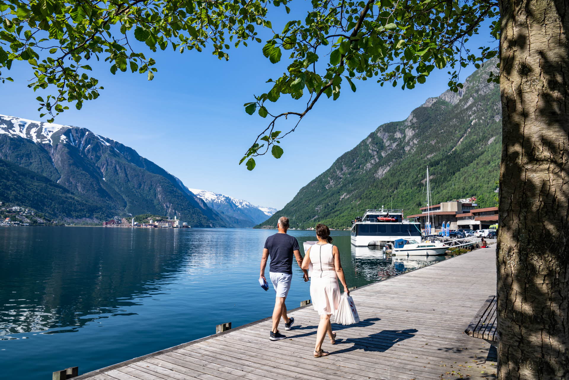 A couple holding hands while walking on a quay by the Hardangerfjord. Birch leaves above, boats in front of them and mountains with snowy peaks in the background.