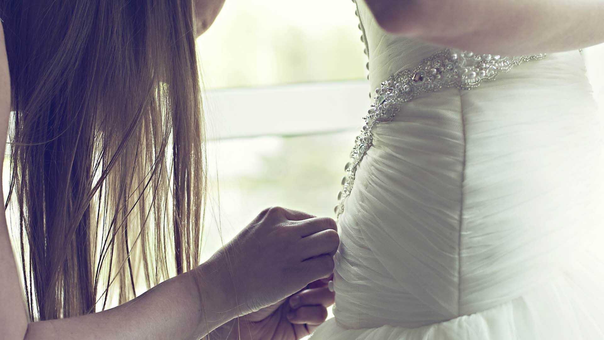 A woman with long hair helps a bride with the back of her wedding dress