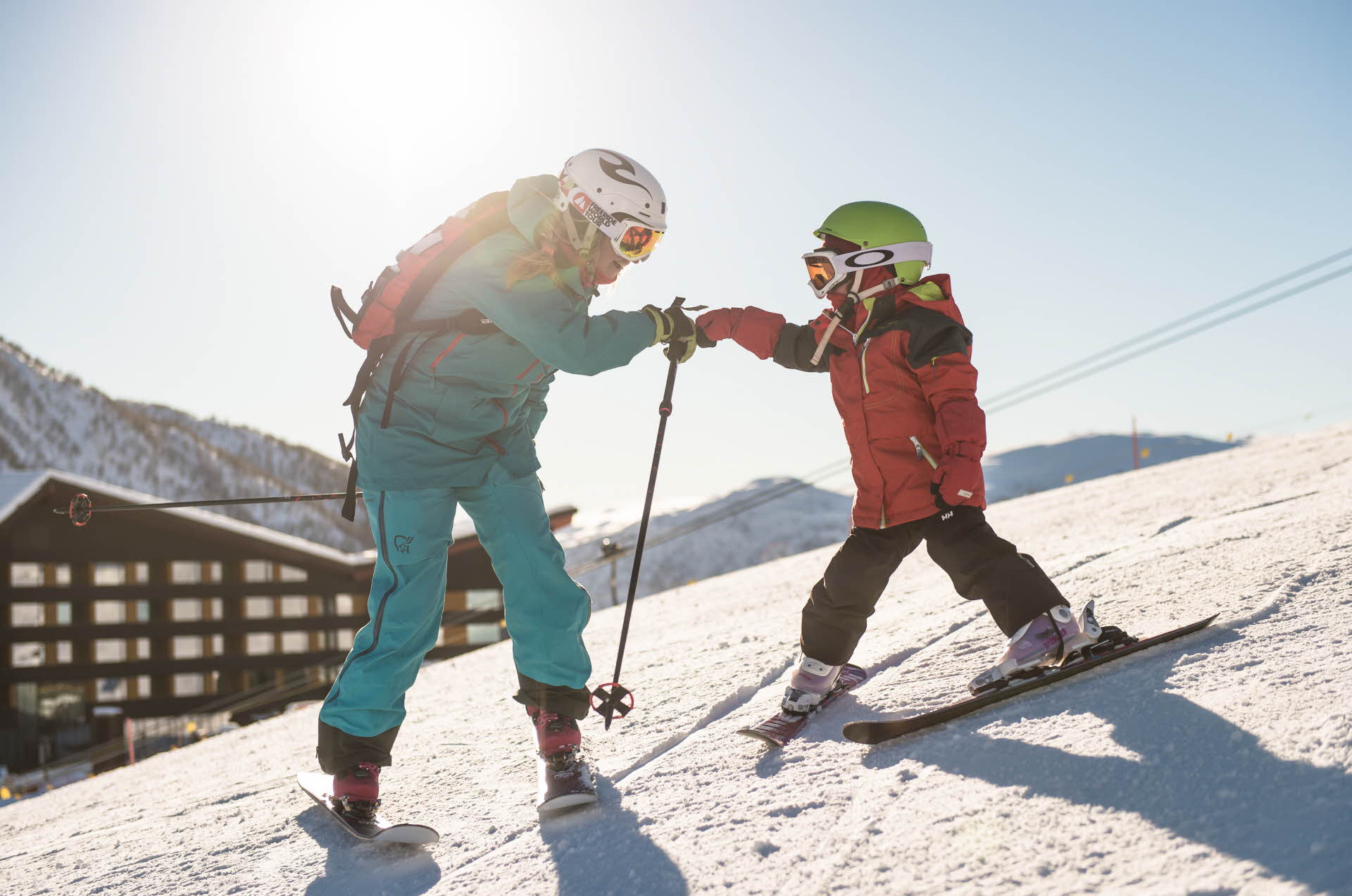 一个大人和一个小孩在滑雪坡上击拳。背景中是迈克达林酒店 (Myrkdalen Hotel)。