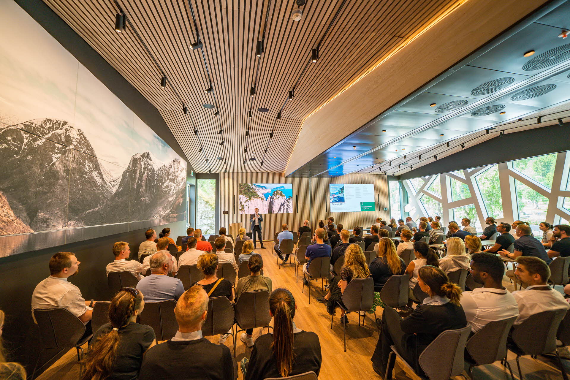 Des personnes assises dans une vaste salle de conférence avec une grande photographie sur l’un des murs, l’autre mur étant occupé par les fenêtres, et un écran au fond.