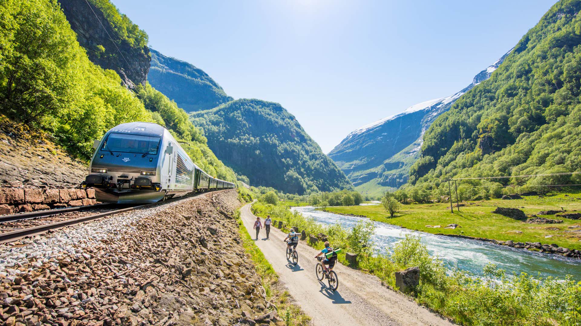 Une journée d’été ensoleillée dans la vallée de Flåm, avec deux cyclistes s’apprêtant à croiser deux randonneurs, entre le train de Flåm et la rivière