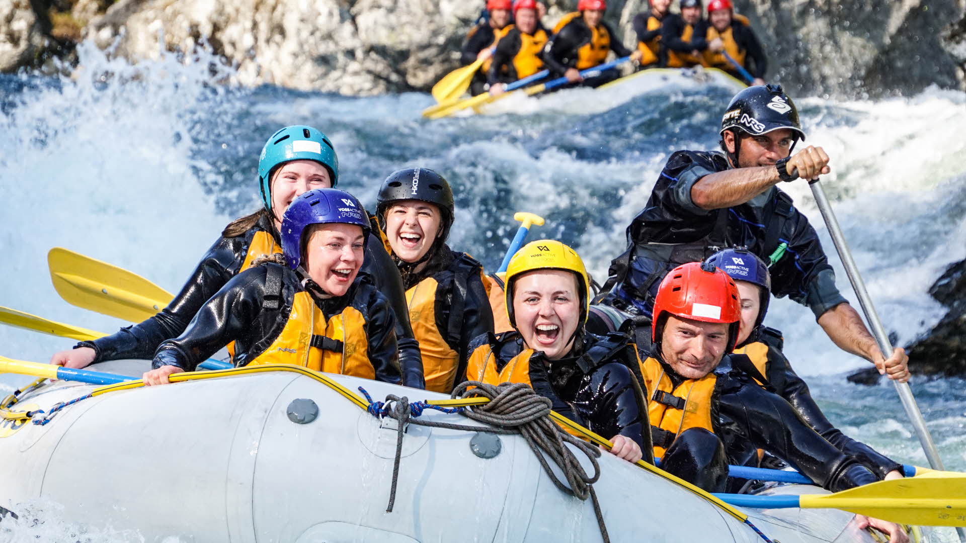 Eine Gruppe von lachenden Menschen in einem Raft bei der Fahrt durch eine Stromschnelle. Im Heck des Bootes der paddelnde Guide, dahinter ein weiteres Boot.