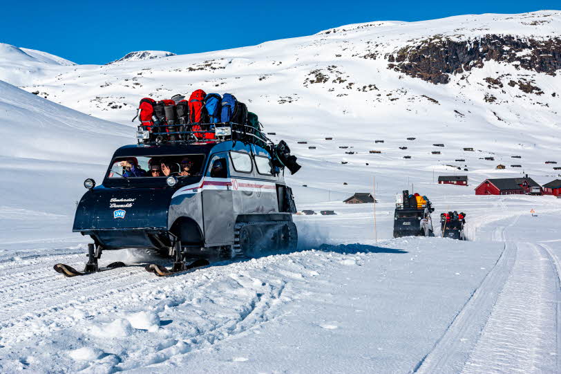 3 dark blue 1960s snow coaches packed with people and gear driving in Jotunheimen winter landscape on a sunny day