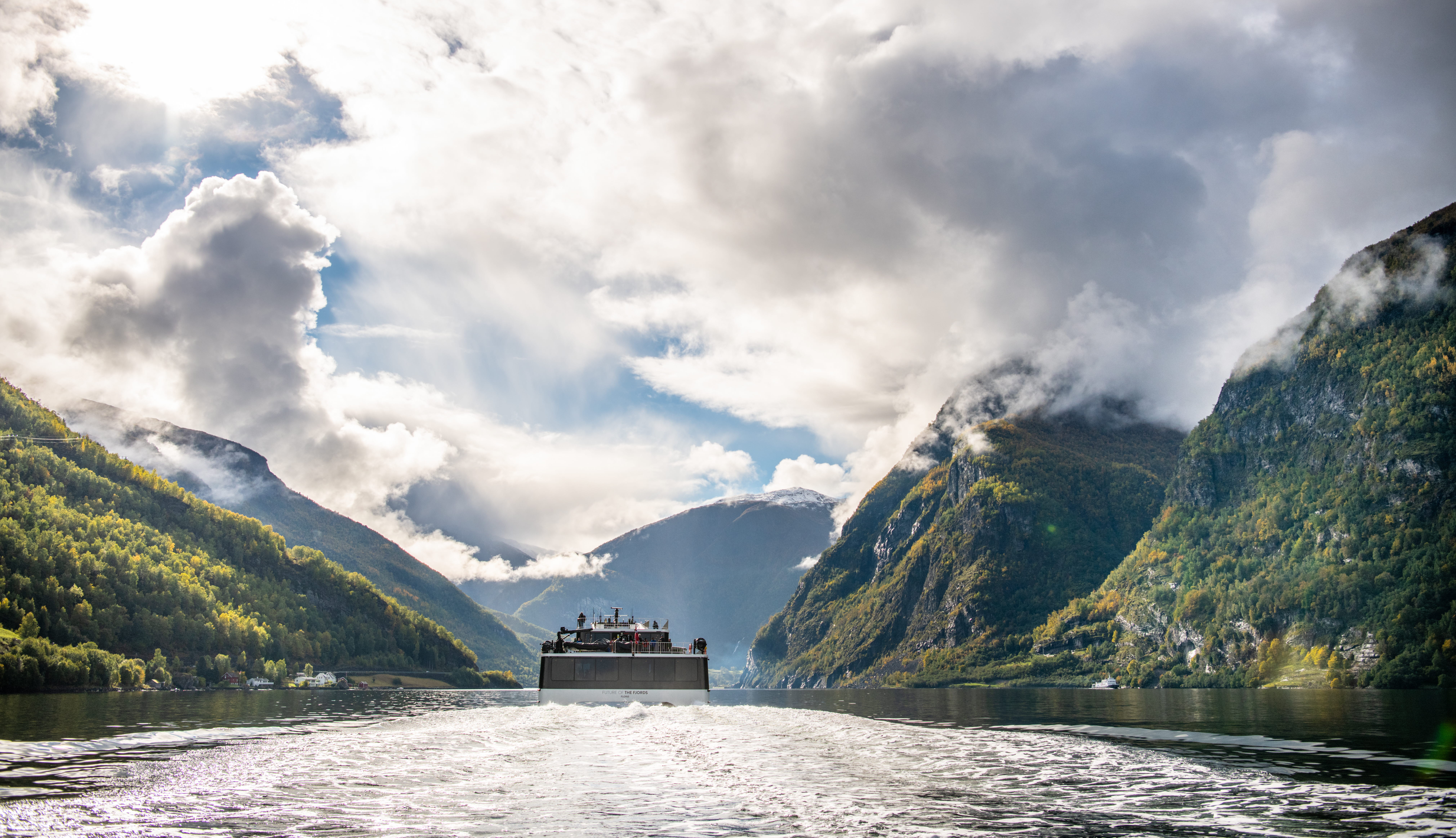 Vue de la poupe d’un catamaran électrique naviguant sur le Nærøyfjord. Montagnes partiellement surmontées de nuages.