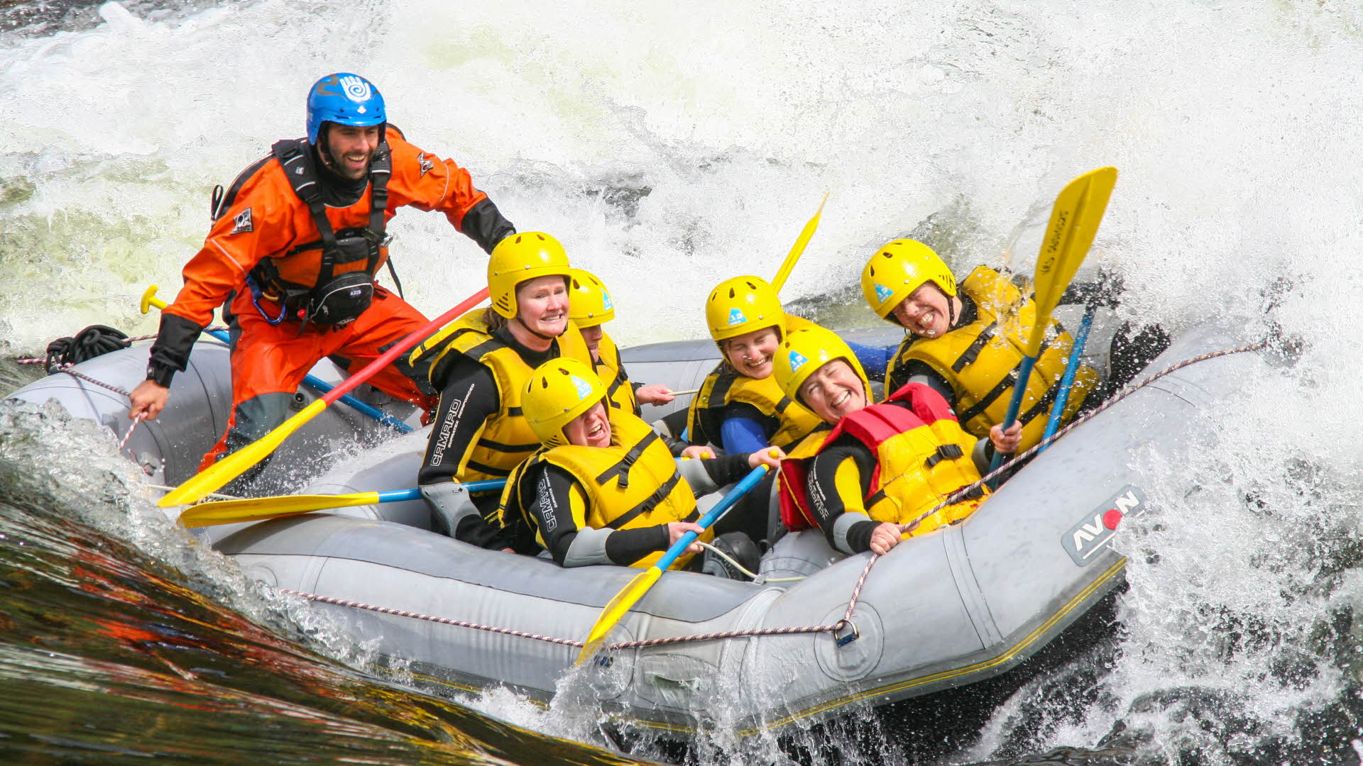 五位头戴黄色头盔身穿救生衣的女士正坐在一艘即将冲入一个巨浪的皮划艇上。一位身穿干式潜水服的导游正站在后面。