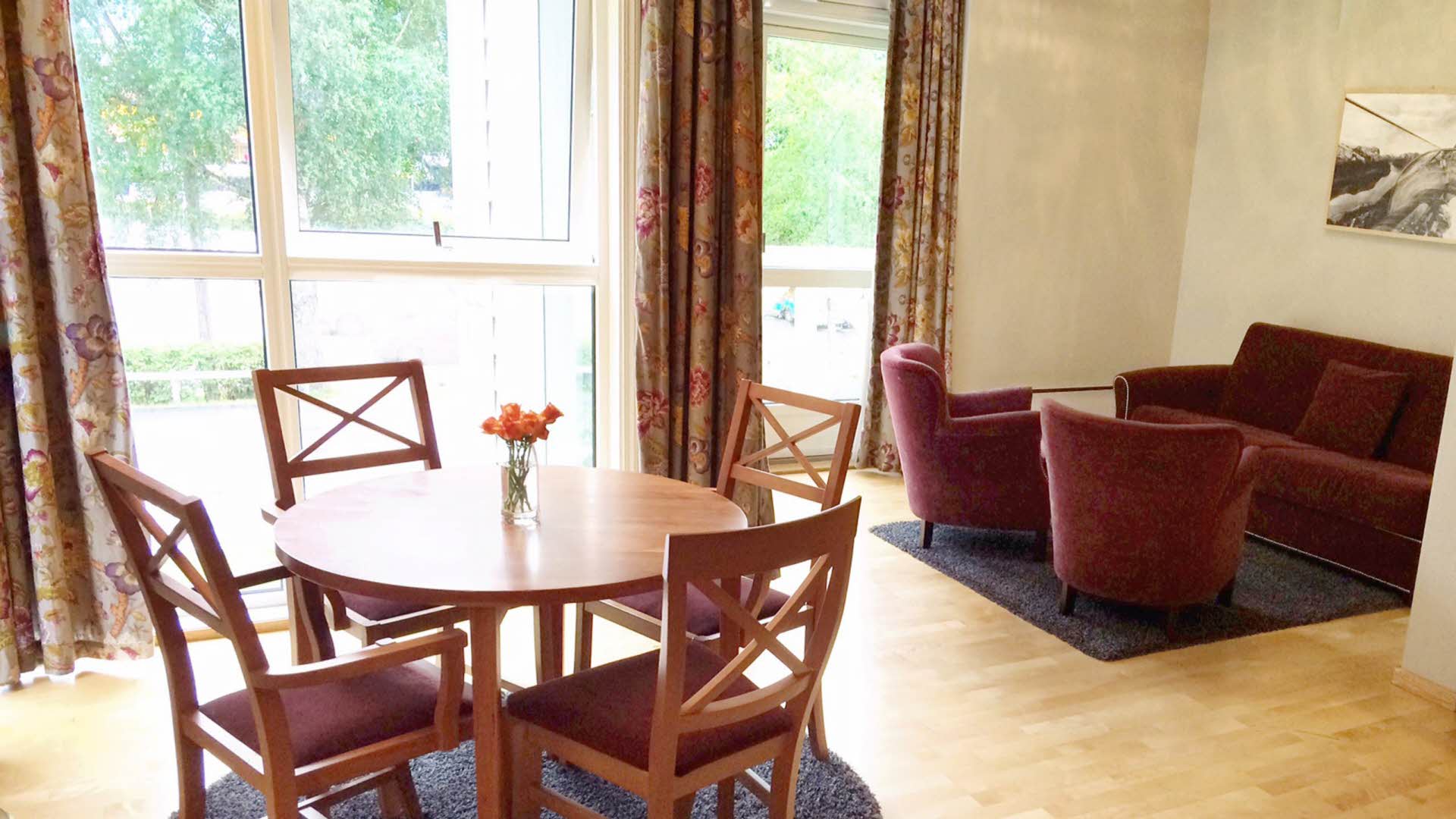 Une table à manger avec quatre chaises, et un coin salon avec deux fauteuils rouges et un canapé rouge devant les grandes fenêtres d’une suite junior du Fretheim Hotel