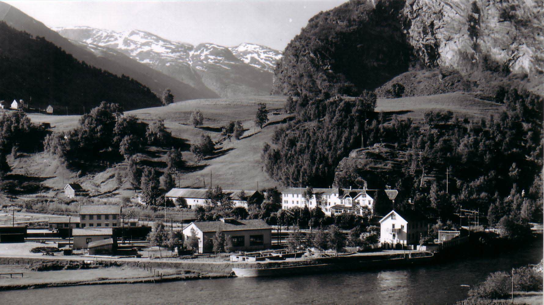 Ein altes Schwarz-Weiß-Foto von Flåm: ein Frachtschiff am Kai, das Hotel Fretheim und der Hügel Fretheimshaugen.