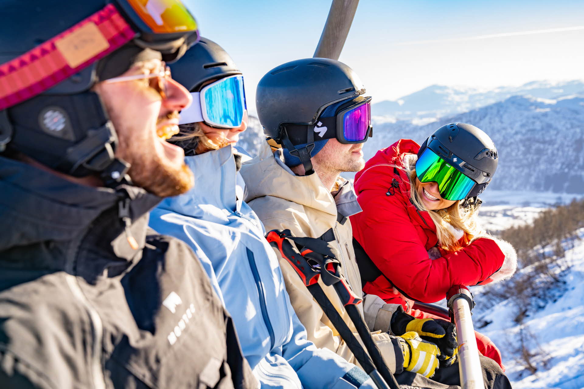 スキー場のリフトに座って笑っているヘルメットとゴーグルを着けた四人。 