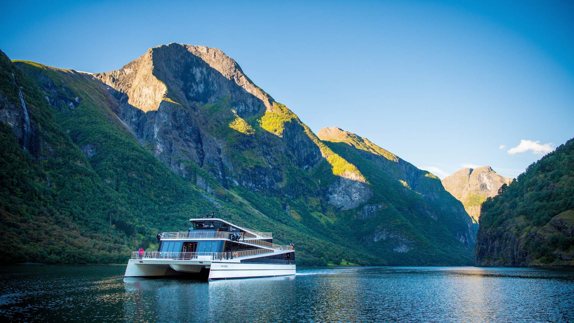 夏にネーロイフィヨルド(Naeroyfjord)を航海するフューチャー オブ ザ フィヨルド号の前方と中央に立つ女性のパノラマビュー