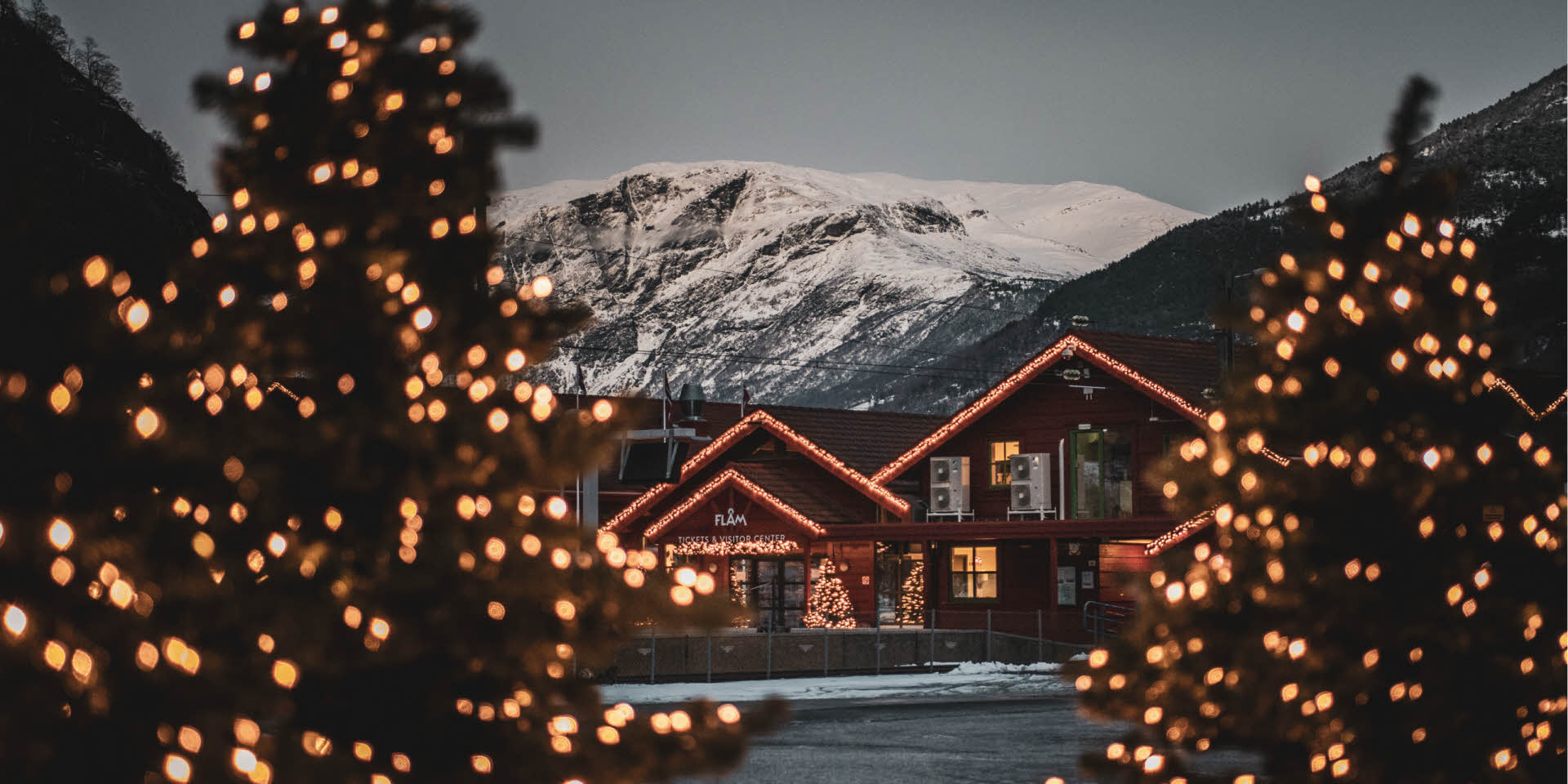 Flåm Visitor Center decorated with Christmas lights framed by Christmas trees and a snowcovered mountain behind. 
