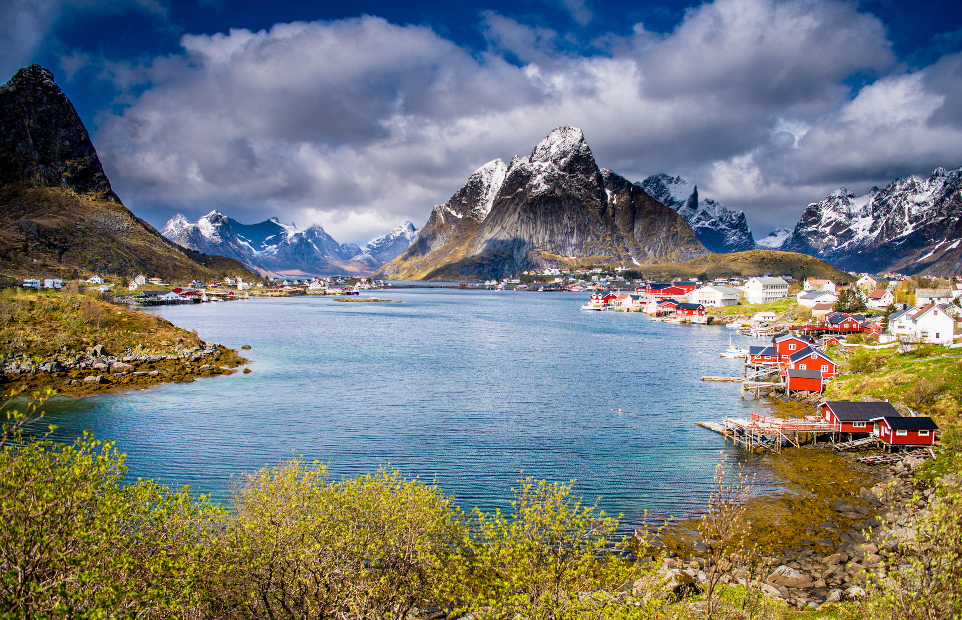 A village by a fjord on the Lofoten Islands and steep, majestic snow-capped peaks. A dramatic cloudy sky.
